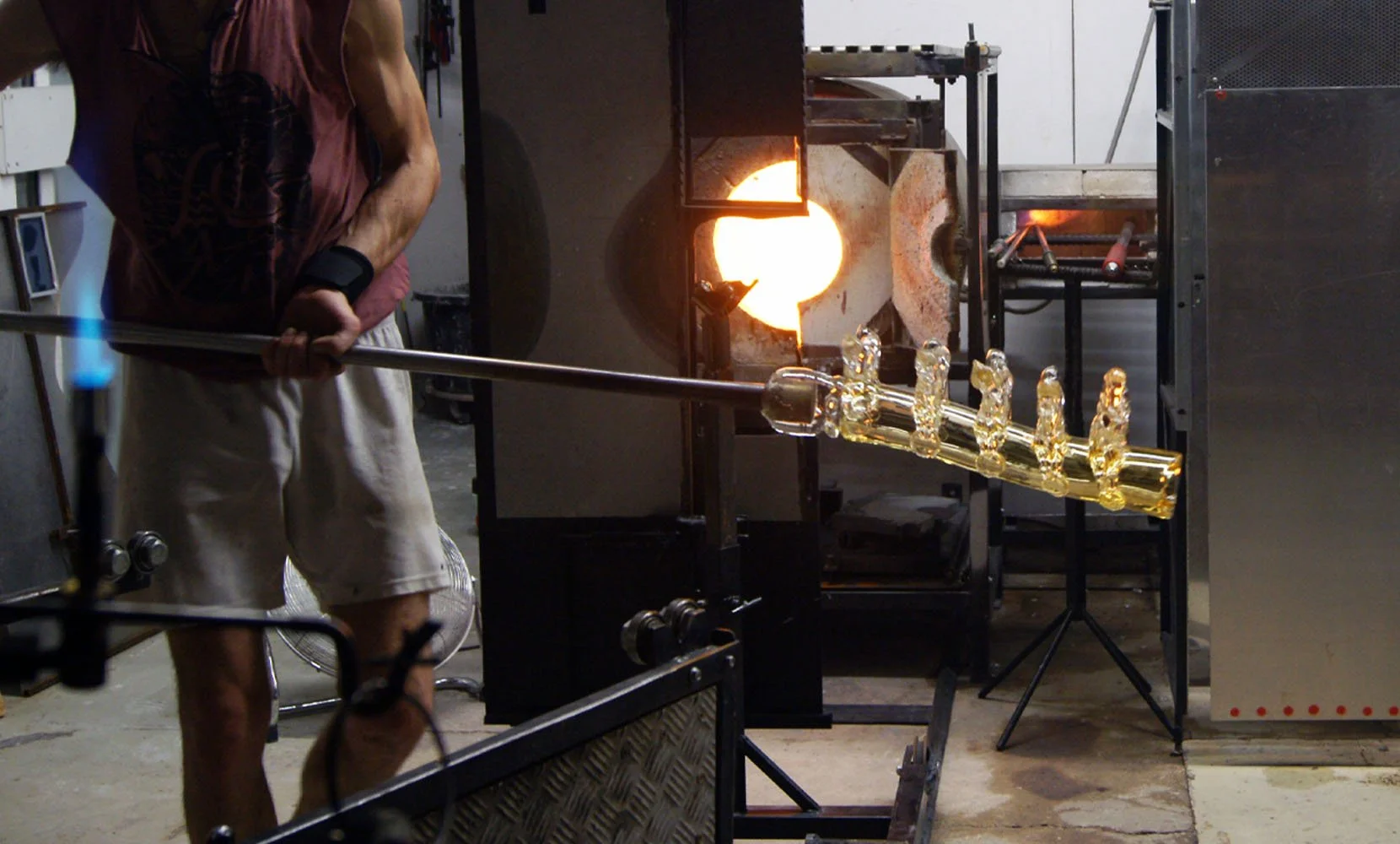 A glassblower shaping molten glass in a workshop, with a furnace glowing in the background.
