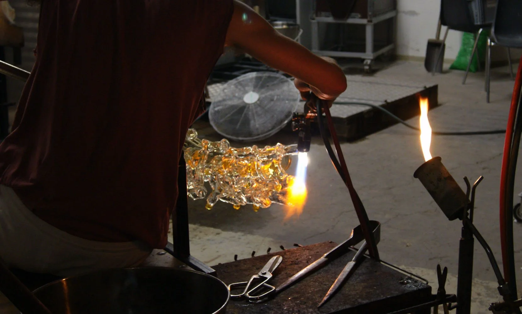 Glassblower shaping molten glass in a workshop, with a blowtorch and tools on the workbench.