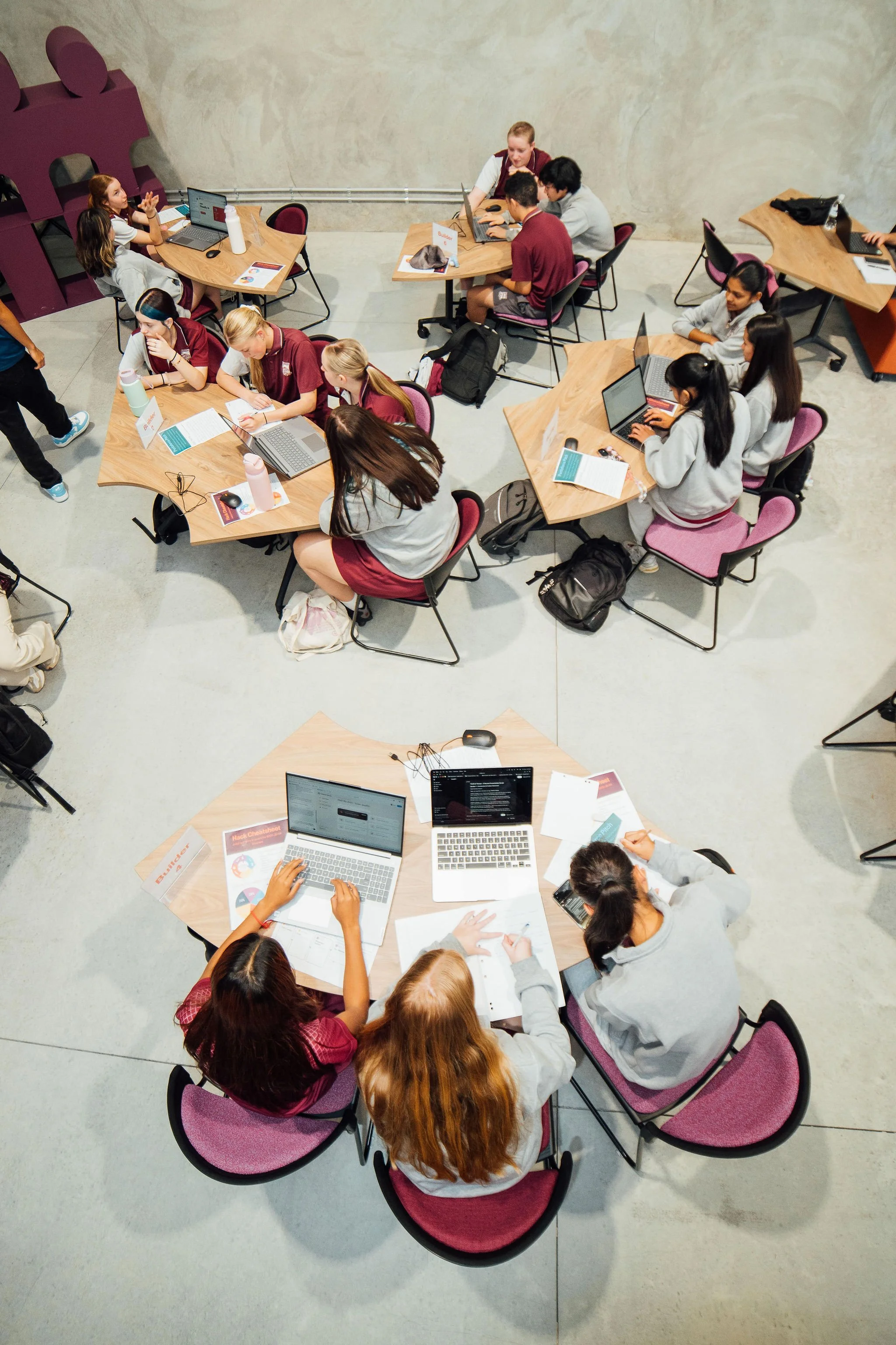 Students gather around tables working on laptops, writing, and discussing in a classroom or workshop setting.