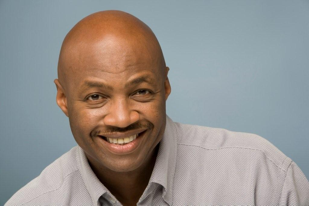 Smiling African American man with a bald head, wearing a light gray dress shirt, against a blue background.