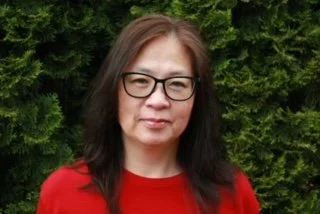 A woman with long dark hair, wearing glasses and a red top, standing outdoors with green foliage in the background.