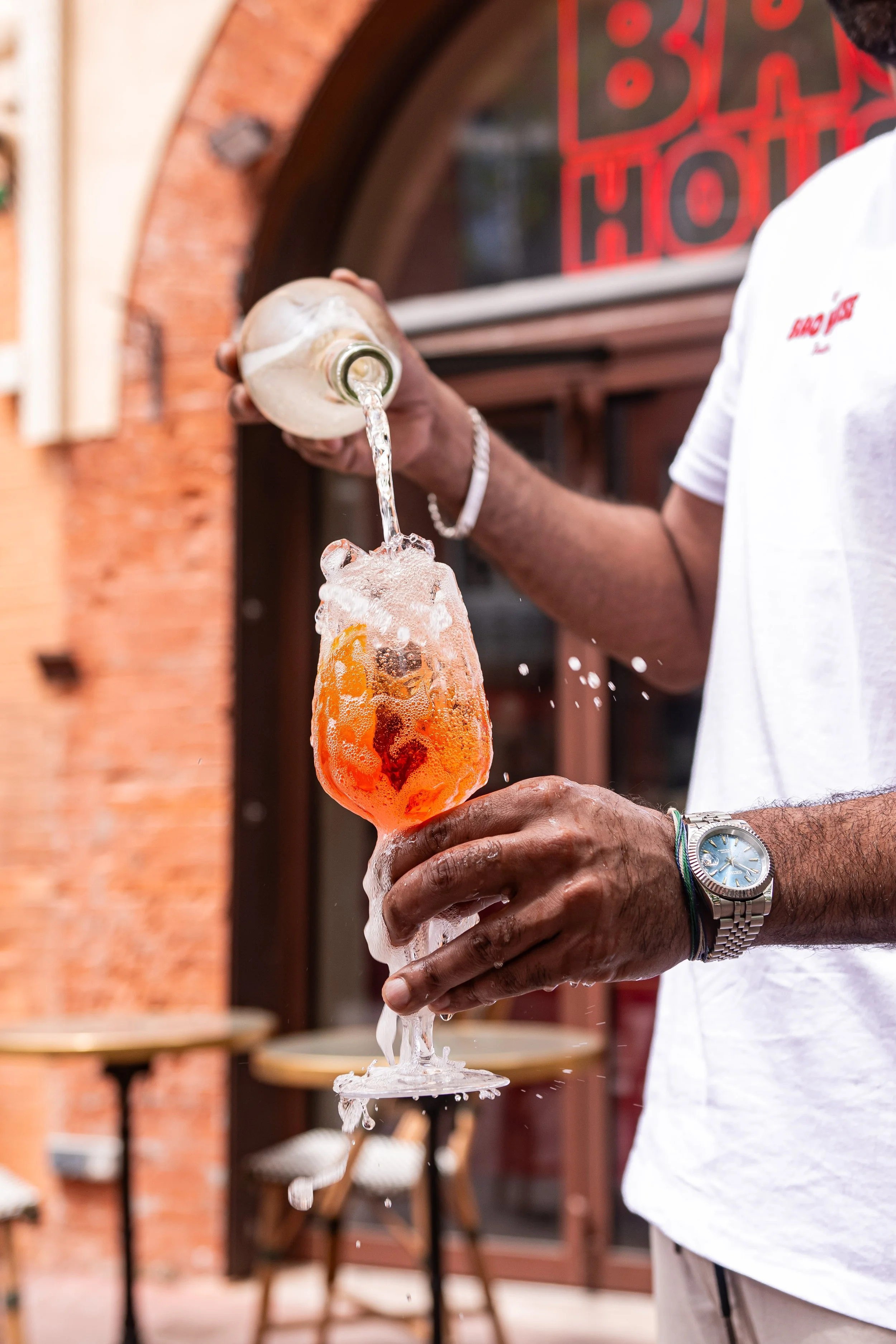 Un homme verse de la crème dans un verre à cocktail lors d'une fête ou d'un événement en extérieur, avec un fond de bâtiment en briques et une enseigne lumineuse rouge.