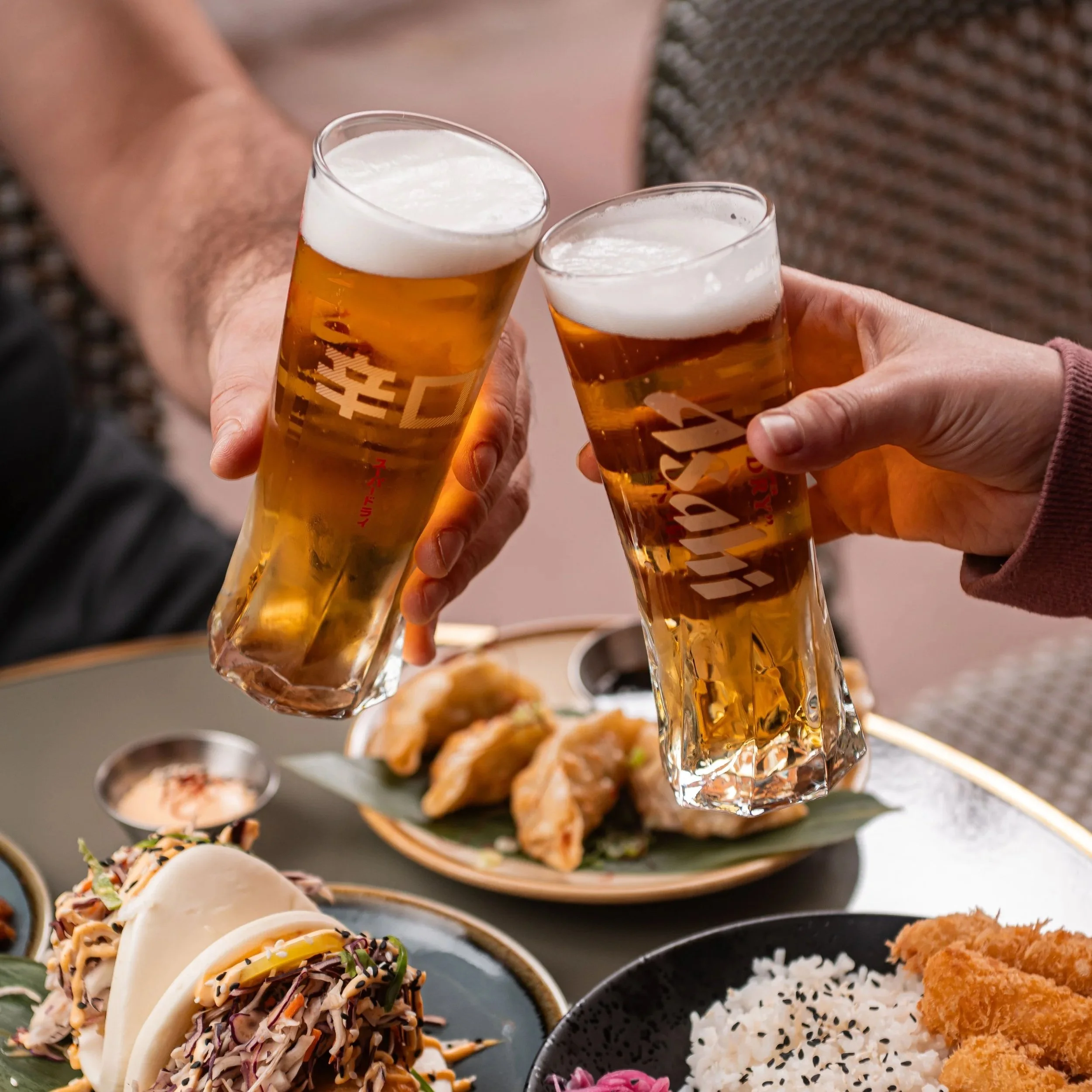 Toasts de bière avec une mousse blanche dense tenus par différentes mains, avec un plateau de sushis et autres plats de nourriture asiatique en arrière-plan.