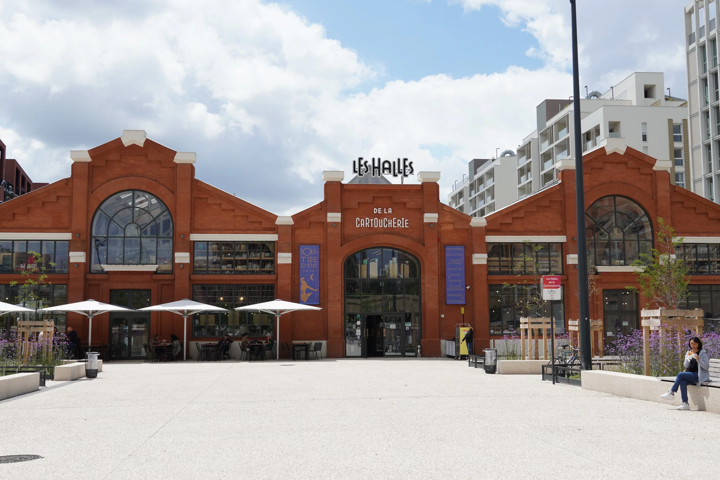 Une façade en briques rouges d'un centre commercial avec un panneau indiquant 'Les Halles de la Cartoucherie'. Il y a des parasols blancs et des personnes assises à l'extérieur, sous un ciel partiellement nuageux.Restaurant Asiatique à Toulouse - BAO