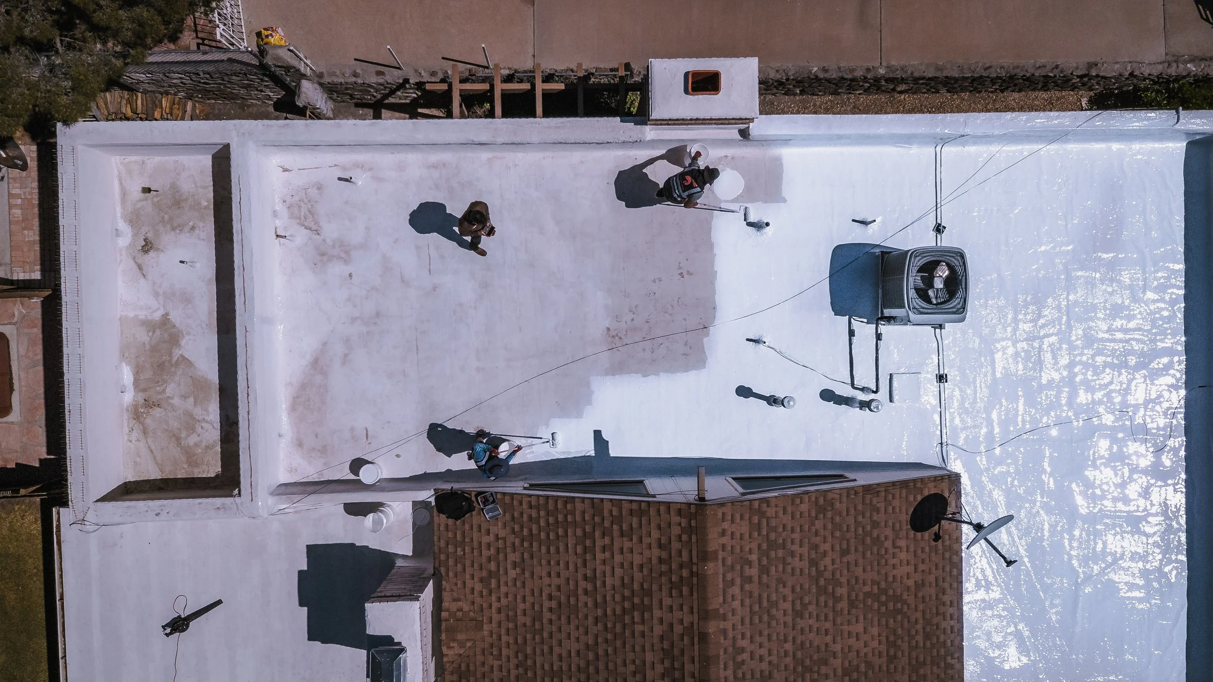 Aerial view of a house roof with multiple textures, shadows, and nearby trees.