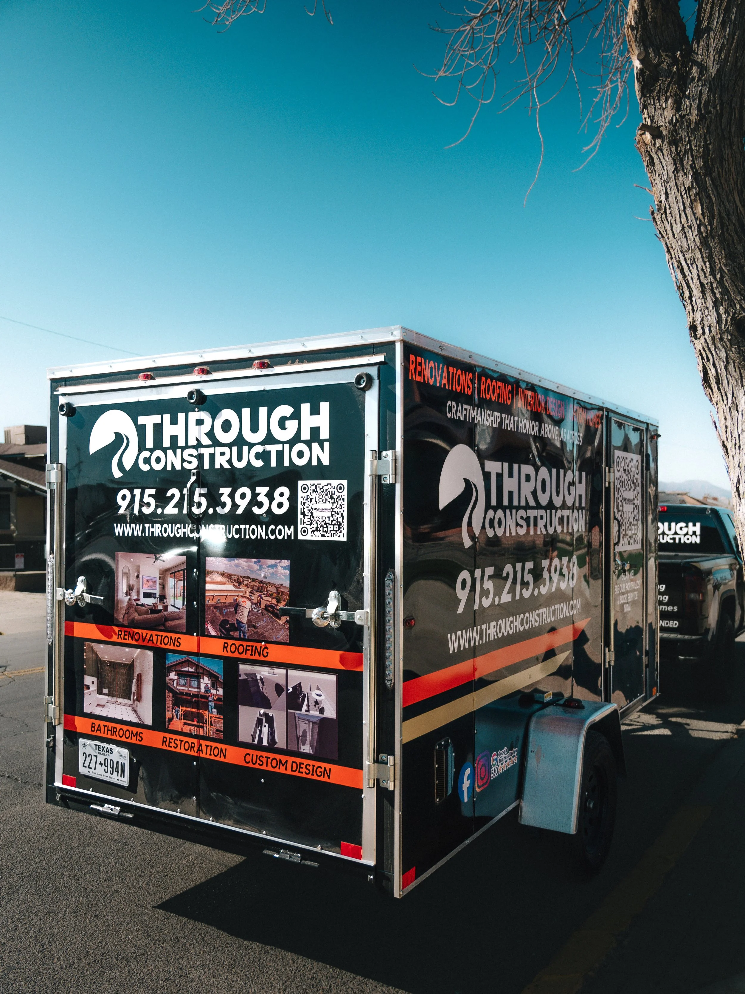A pickup truck parked in front of a house with construction materials in its bed and a worker sitting on a bench nearby. The truck has logos and contact information for a construction company named 'Through Construction.' The house in the background is under renovation, with visible roofing work.
