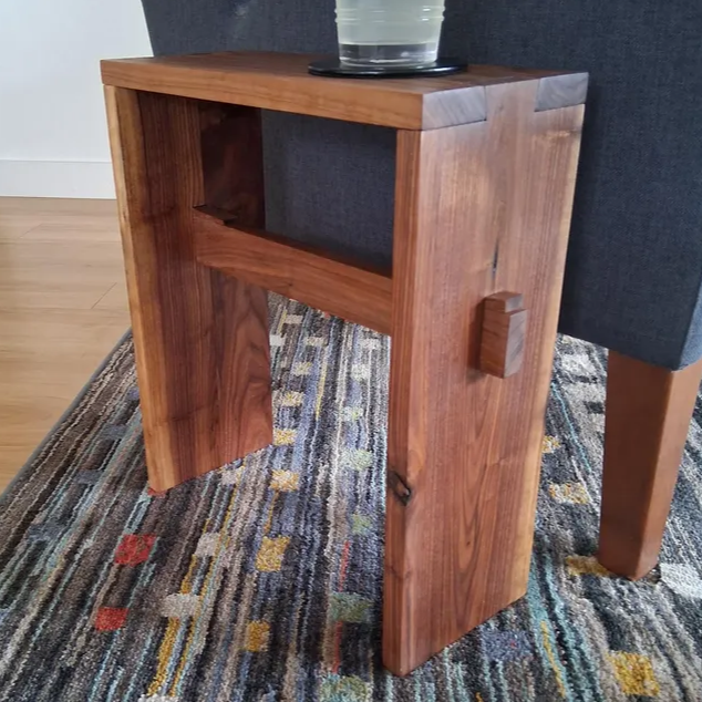 Small wooden side table with a glass of water on top, placed next to a dark gray upholstered chair on a patterned rug.
