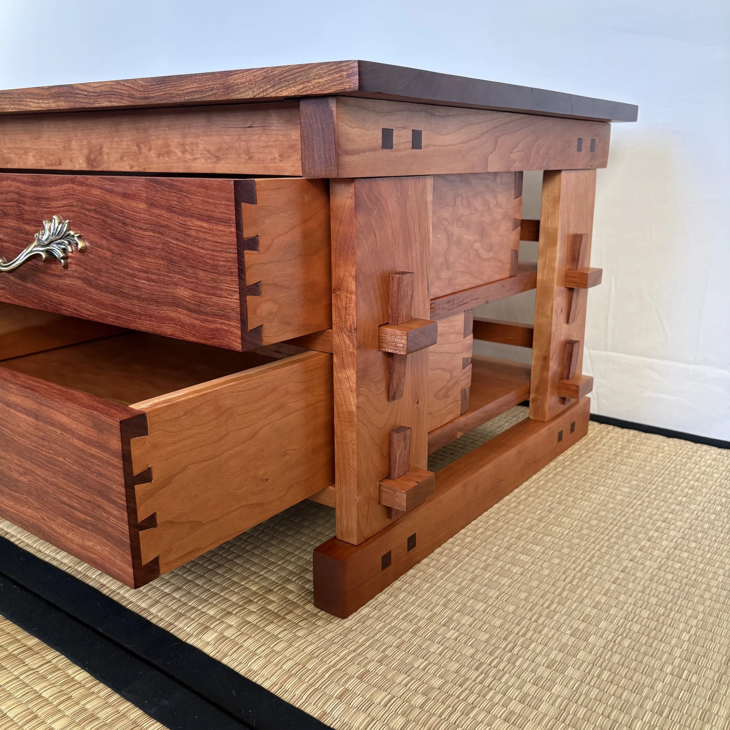 Close-up of a wooden furniture piece, possibly a dresser, with dovetail joints and a decorative silver handle, placed on a woven rug.