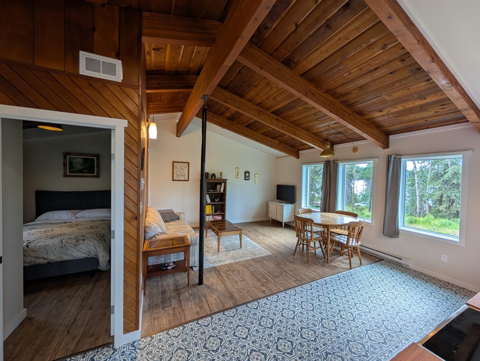Living room with ceiling beams, large windows with curtains overlooking trees, a dining table with chairs, a bookshelf, a small TV stand, and a bedroom with a bed partially visible through a doorway. The room features wood paneling and hardwood floors.