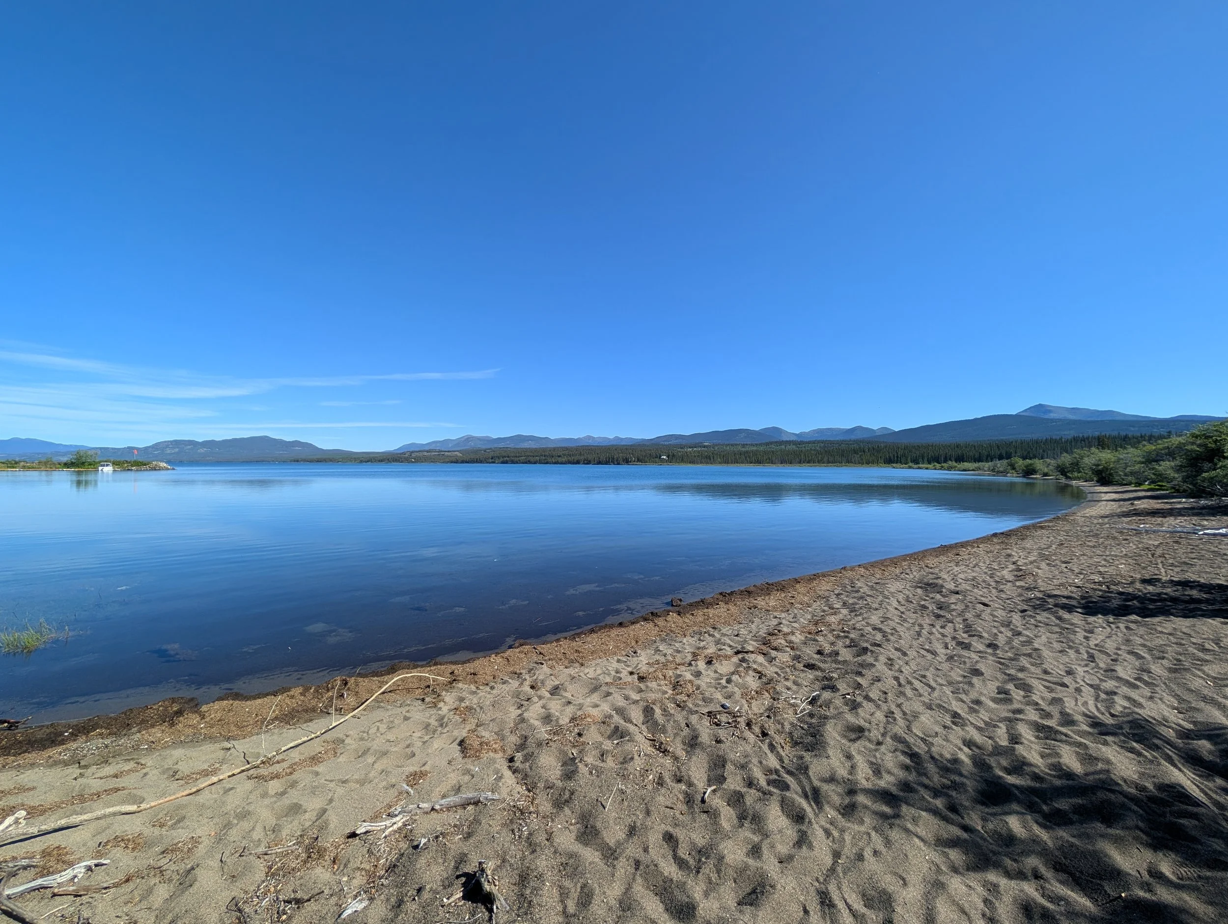Sandy beach, clear blue water, with mountains and a small marina in the background.