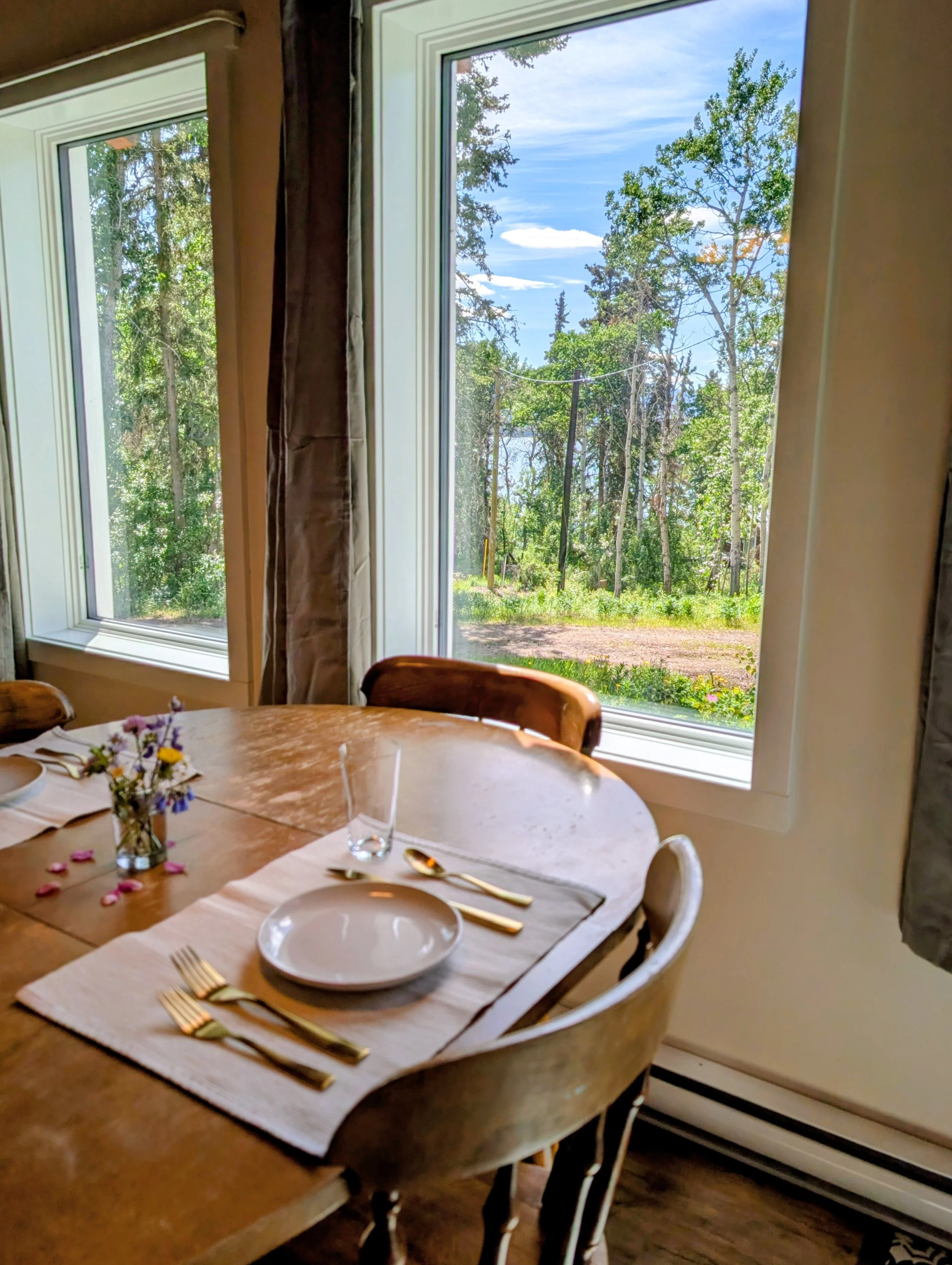Interior of a dining room with a round wooden table set with white plates, gold utensils, clear glasses, and a floral centerpiece. Large windows reveal a scenic view of trees and a blue sky outside.