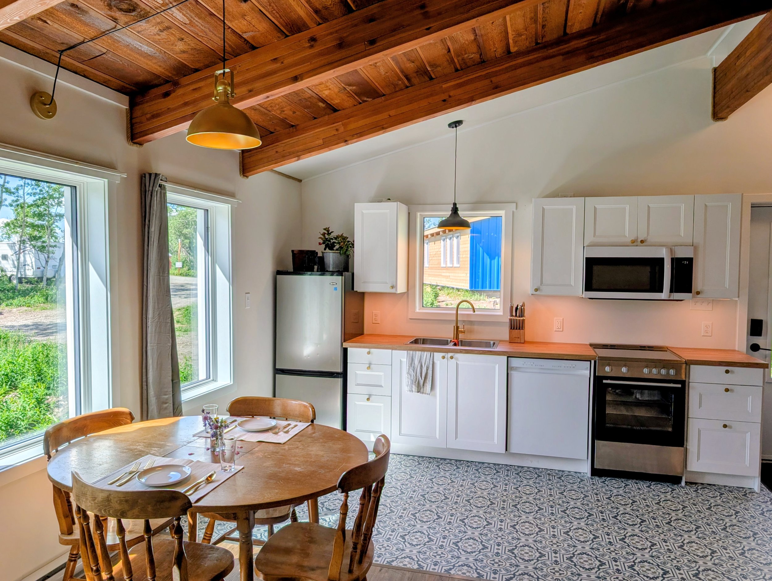 A cozy kitchen and dining area with a wooden beamed ceiling, white cabinets, a small window, a refrigerator, oven, and a round wooden dining table with chairs set for a meal.