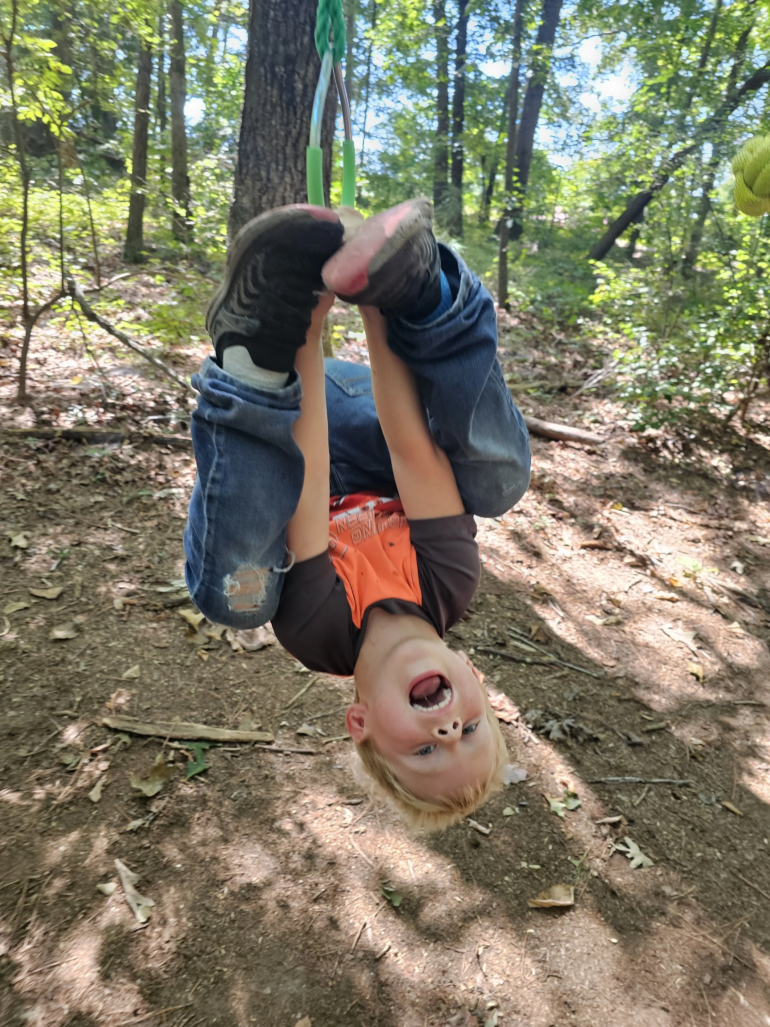 A young boy hanging upside down from a tree branch in a wooded forest, holding onto a green and blue rope, with a playful expression on his face.