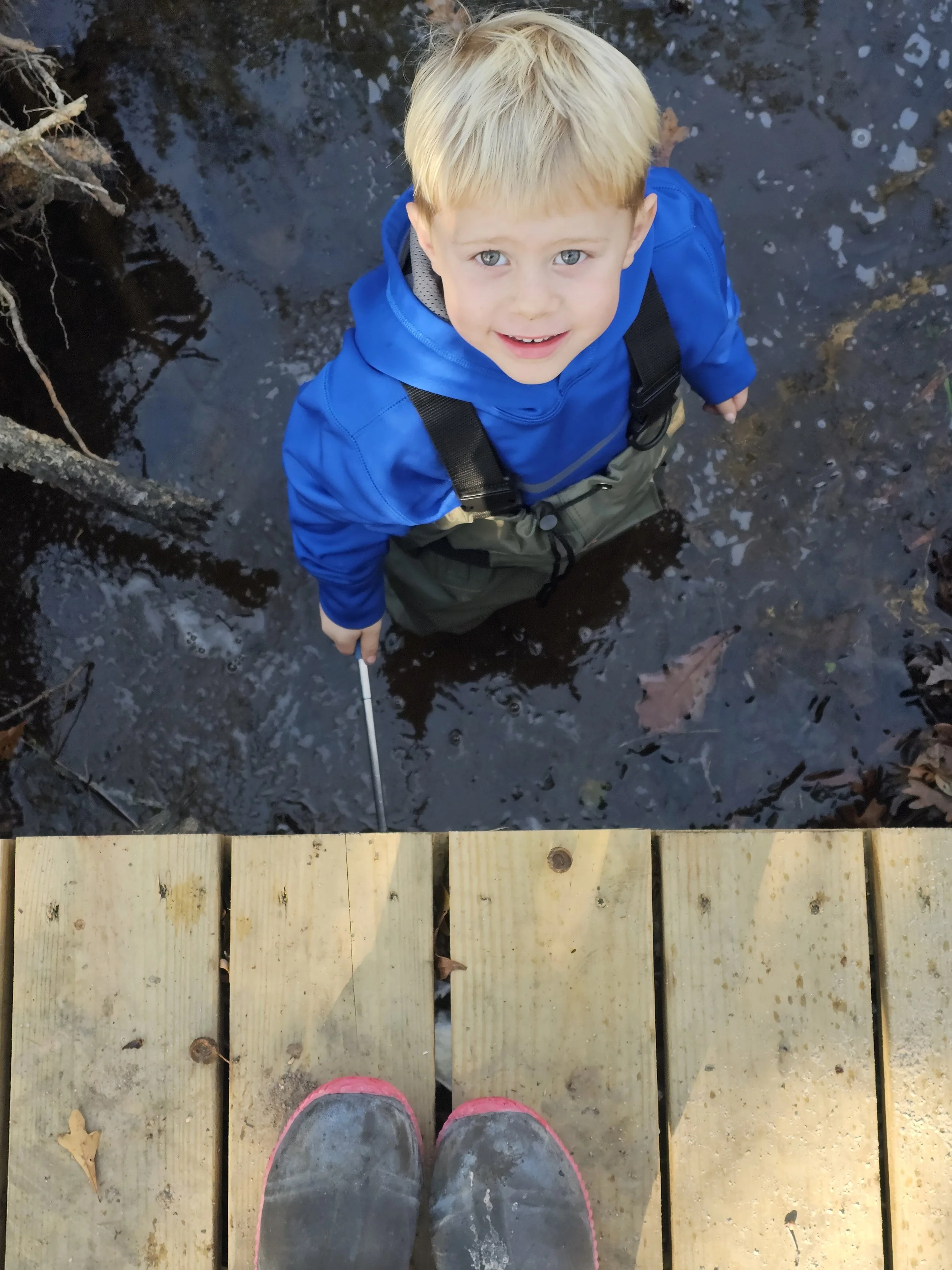 A young boy with blonde hair and blue eyes standing in a shallow creek, looking up at the camera. He is wearing a bright blue jacket and green waterproof pants. The photo is taken from a wooden dock, with a pair of muddy shoes visible at the bottom o