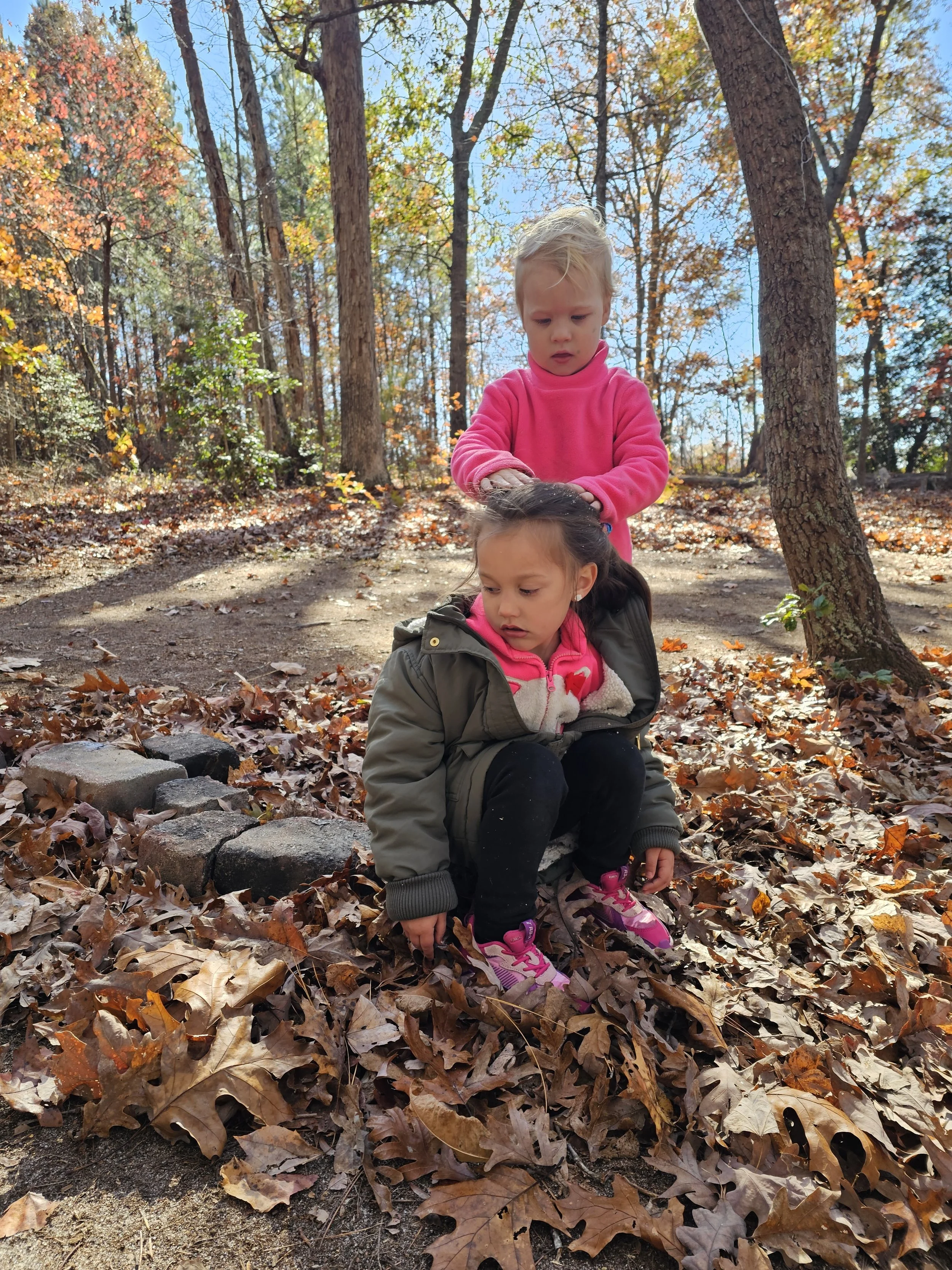 Two young girls playing outdoors on a bed of fallen autumn leaves in a wooded area, with one girl sitting and the other standing behind her, on a small stone bench, surrounded by trees with colorful leaves and clear blue sky.