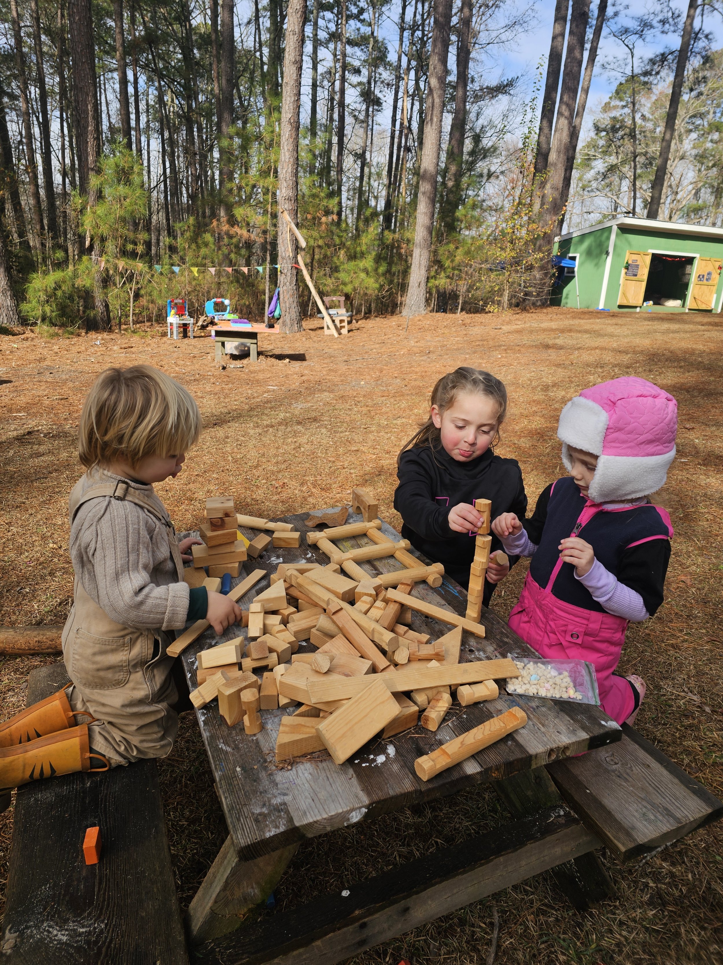 Three children playing outside at a wooden picnic table with wooden blocks, surrounded by trees and a small green shed in the background.