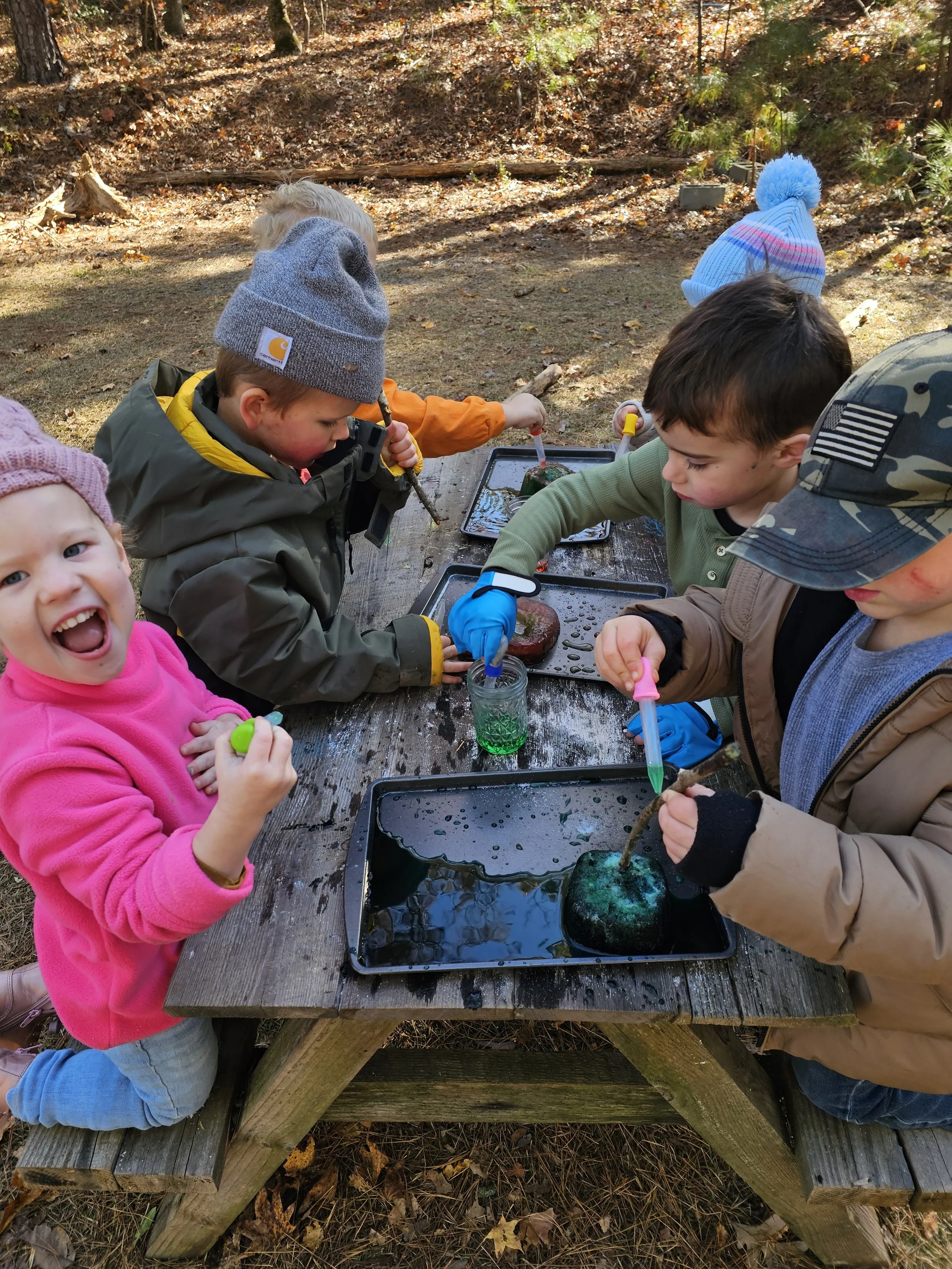 Children gathered around a wooden picnic table outdoors, engaging in an activity with water and colorful pipettes, with autumn trees in the background.