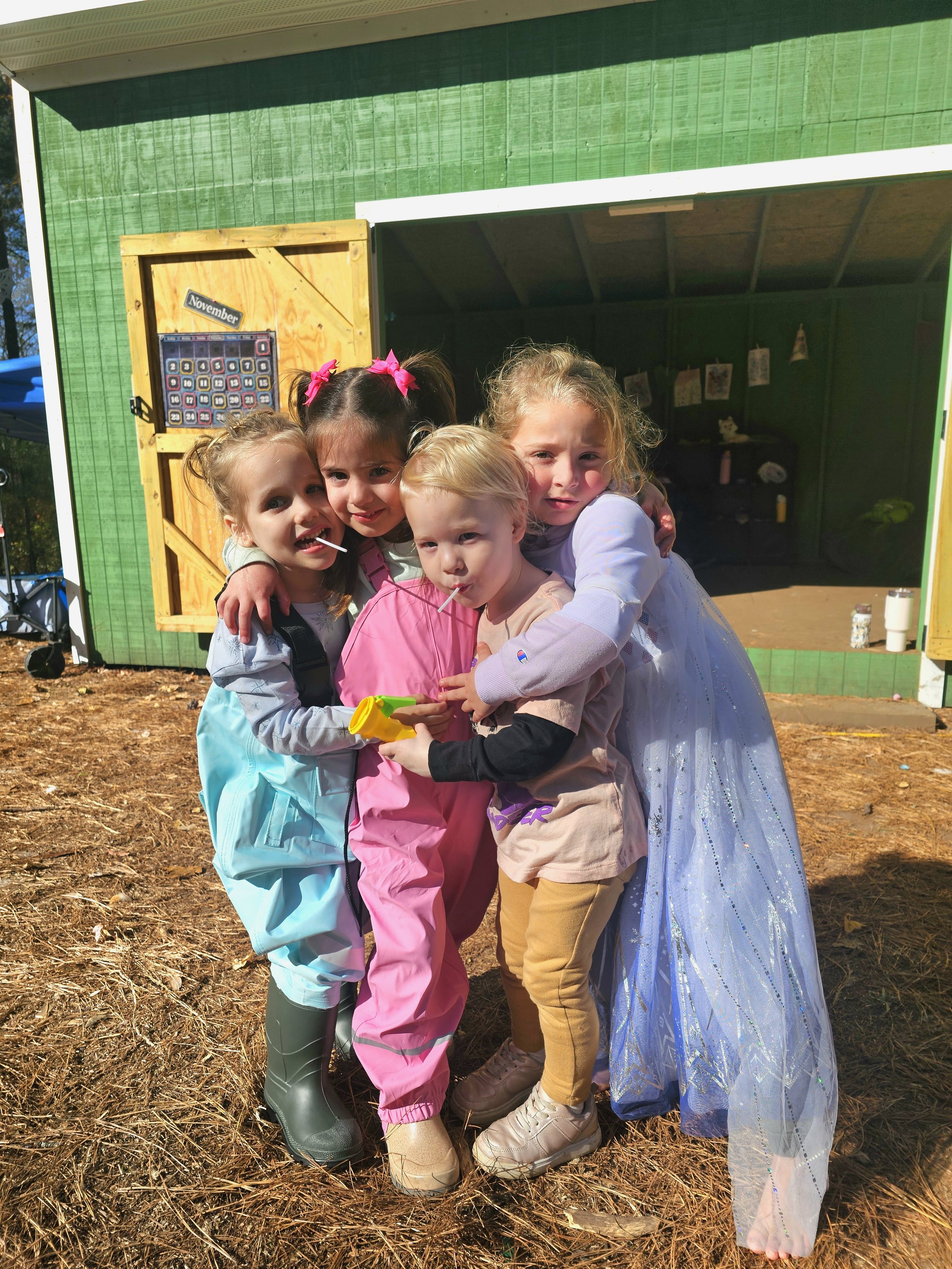 Four young girls dressed in costumes hugging and standing together outside a green wooden playhouse, with autumn leaves on the ground.