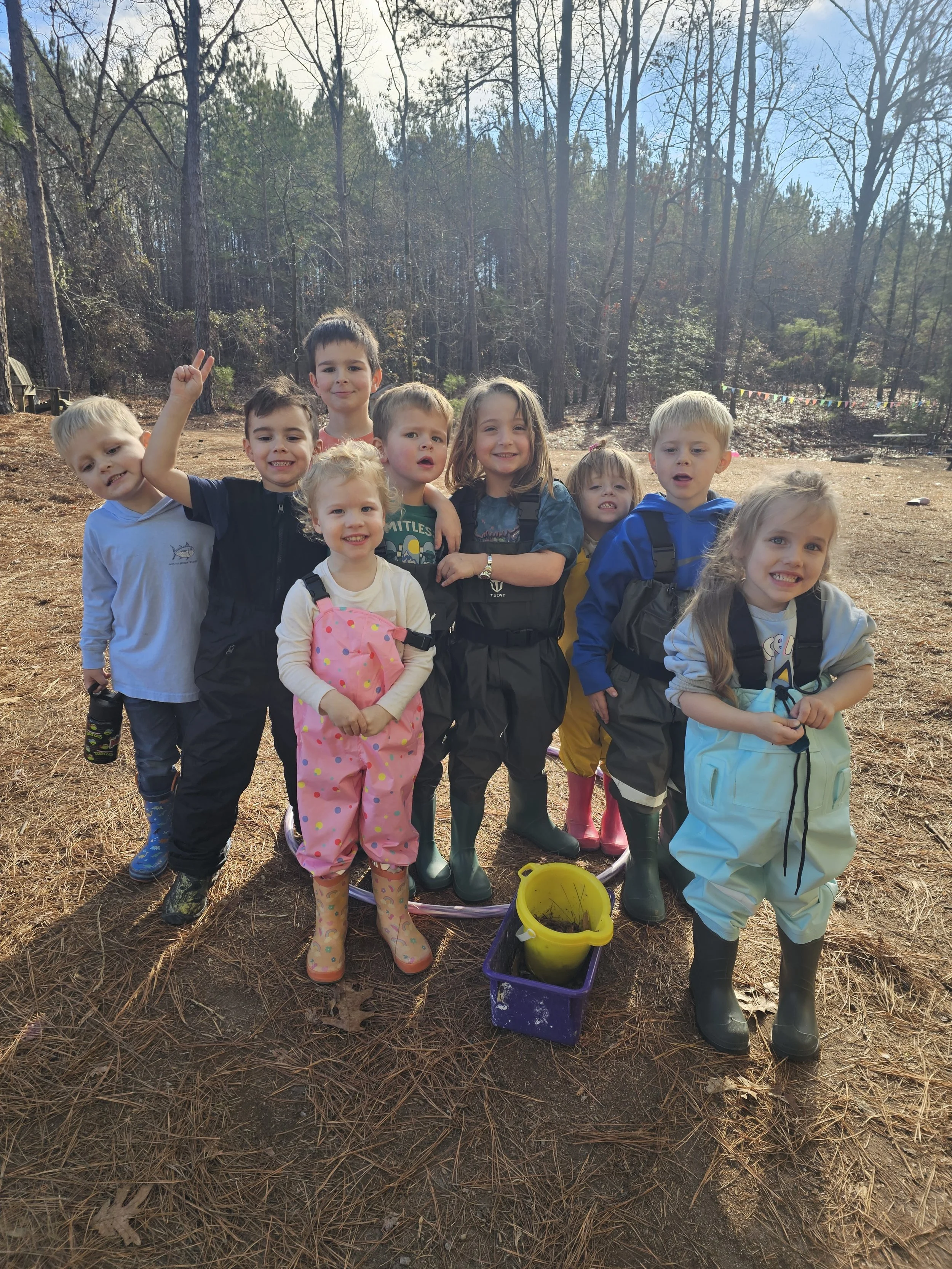 Children dressed in waterproof clothing and boots standing outdoors on a pine needle-covered ground, with trees in the background, posing for a group photo during a outdoor activity.