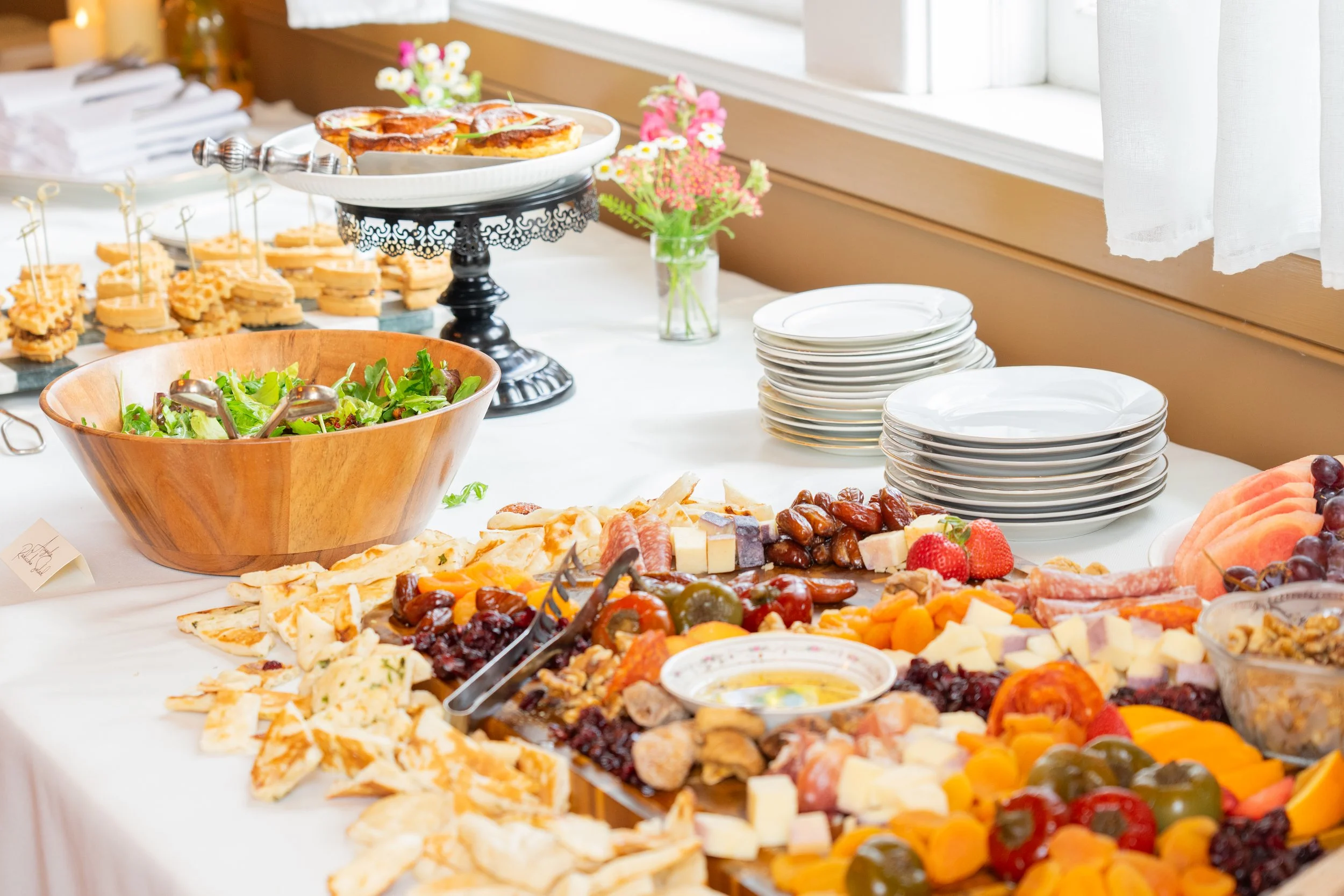 A breakfast or brunch buffet table with plates, a large salad bowl, various cheeses, fresh fruits, baked goods, and small pastries, set near a window with white curtains and floral decorations.