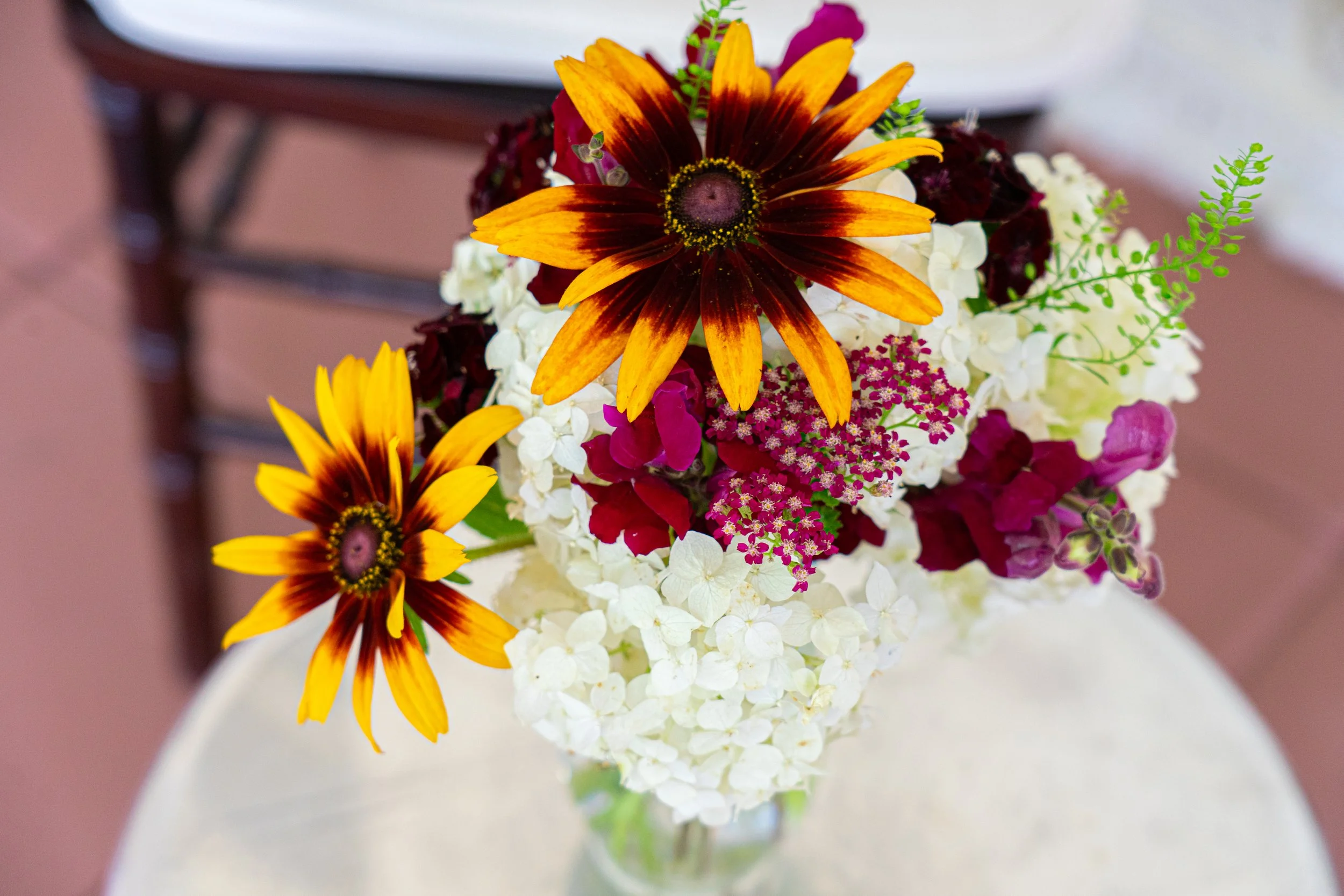 Colorful bouquet of flowers including orange and dark red daisies, white hydrangeas, and purple and pink flowers arranged in a vase on a table.