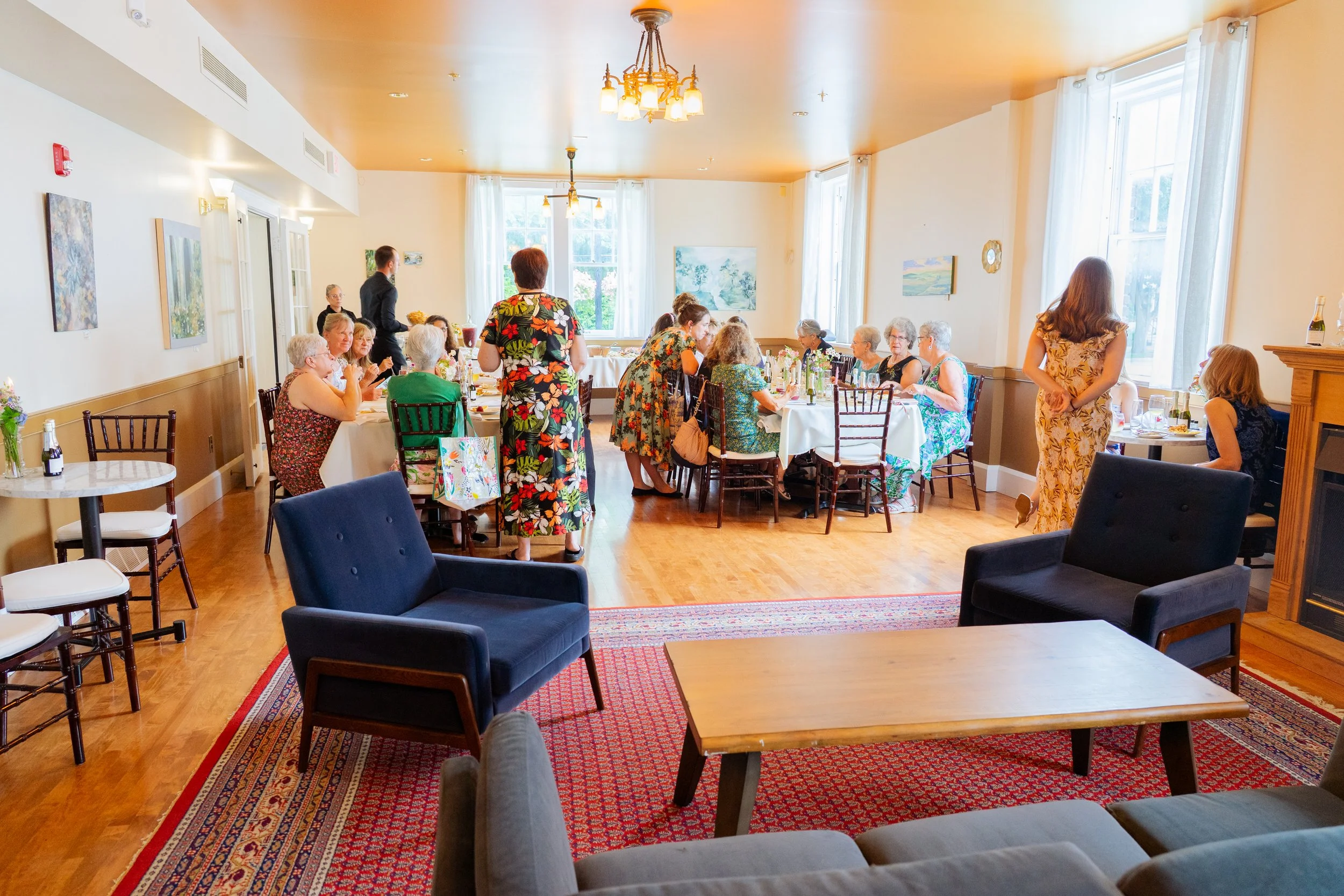 A group of women gathered around dining tables in a bright, spacious dining room, with some standing and some sitting, engaging in conversation. The room has wooden floors, large windows with white curtains, and artwork on the walls.