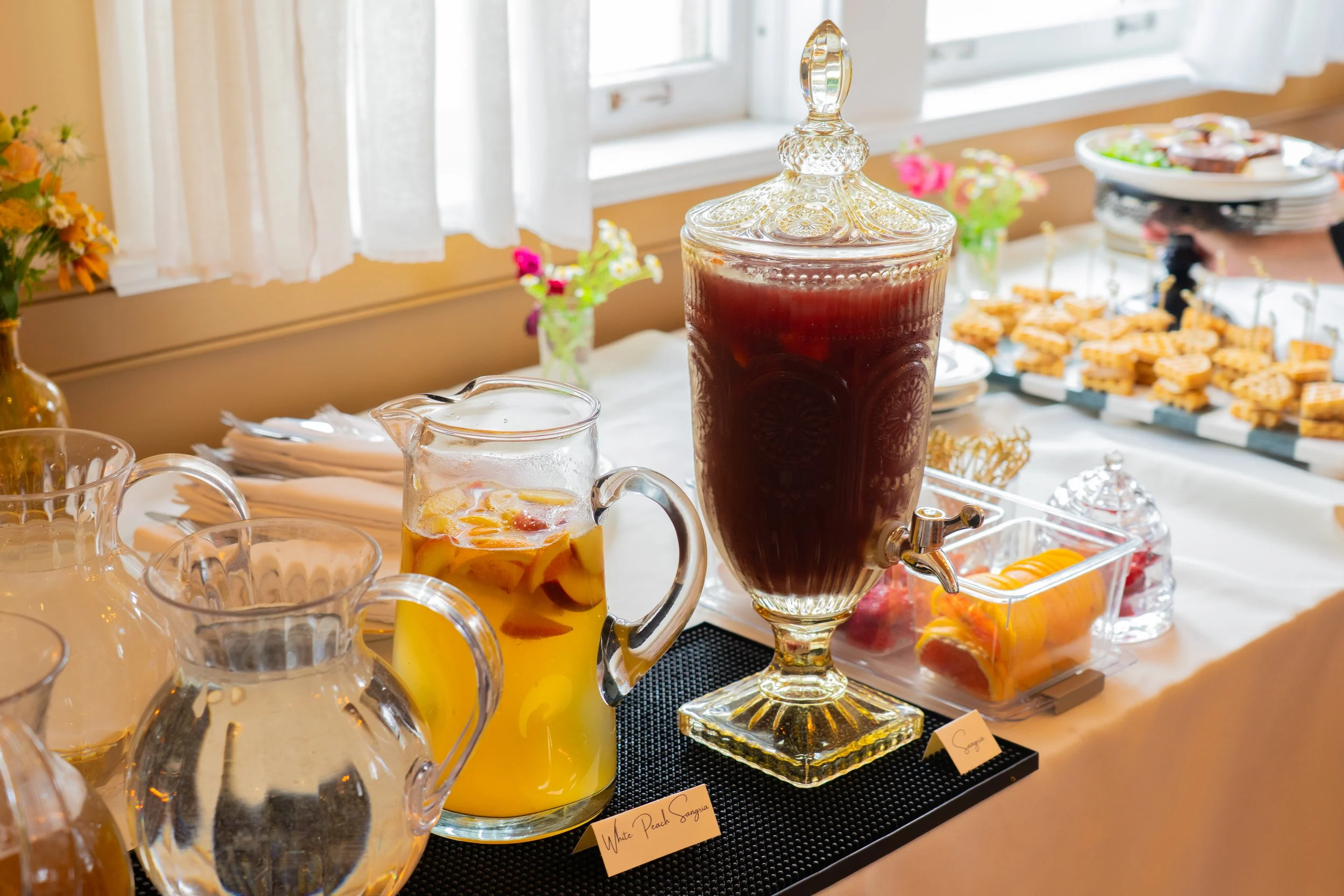 Beverage station with a large glass dispenser of sangria, a pitcher of peach sangria, and a tray of assorted finger foods on a white banquet table near a window with white curtains.