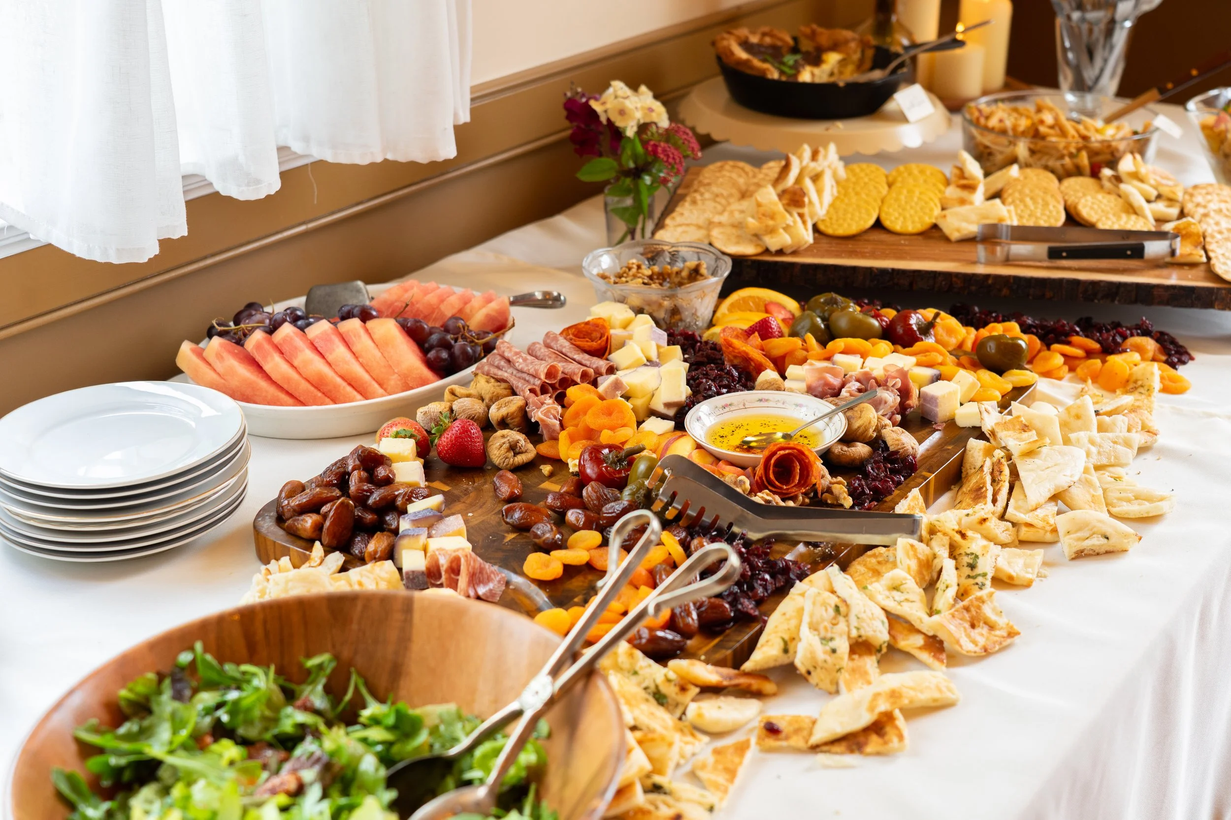 A buffet table with assorted fruits, cheeses, crackers, nuts, dried fruits, and a salad, arranged for a gathering or party.