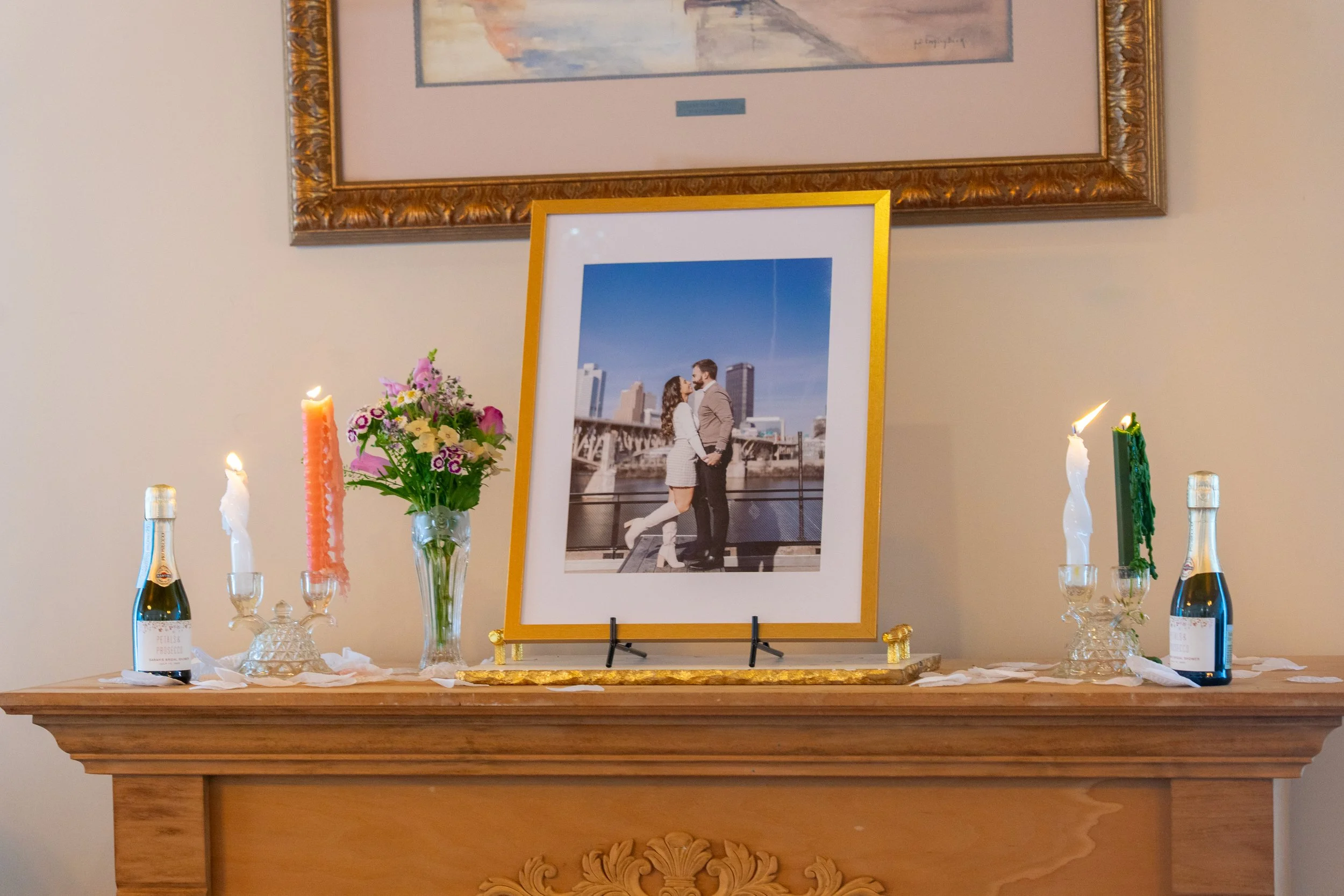 A framed photograph of a couple standing on a bridge in front of a city skyline, placed on a fireplace mantel decorated with candles, flowers, and champagne bottles.