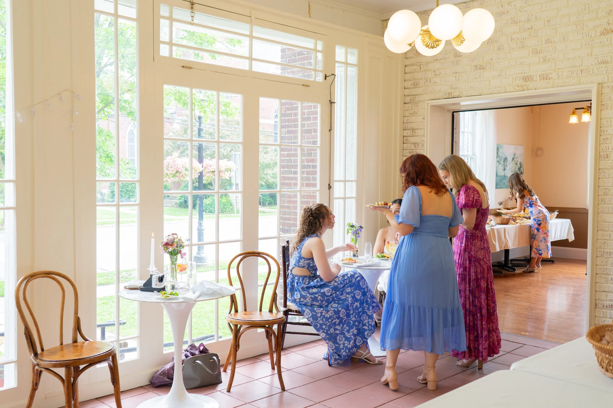 A group of women gathered for a meal in a bright, sunlit dining area with large windows, decorated for a celebration or brunch, with food and floral arrangements visible.