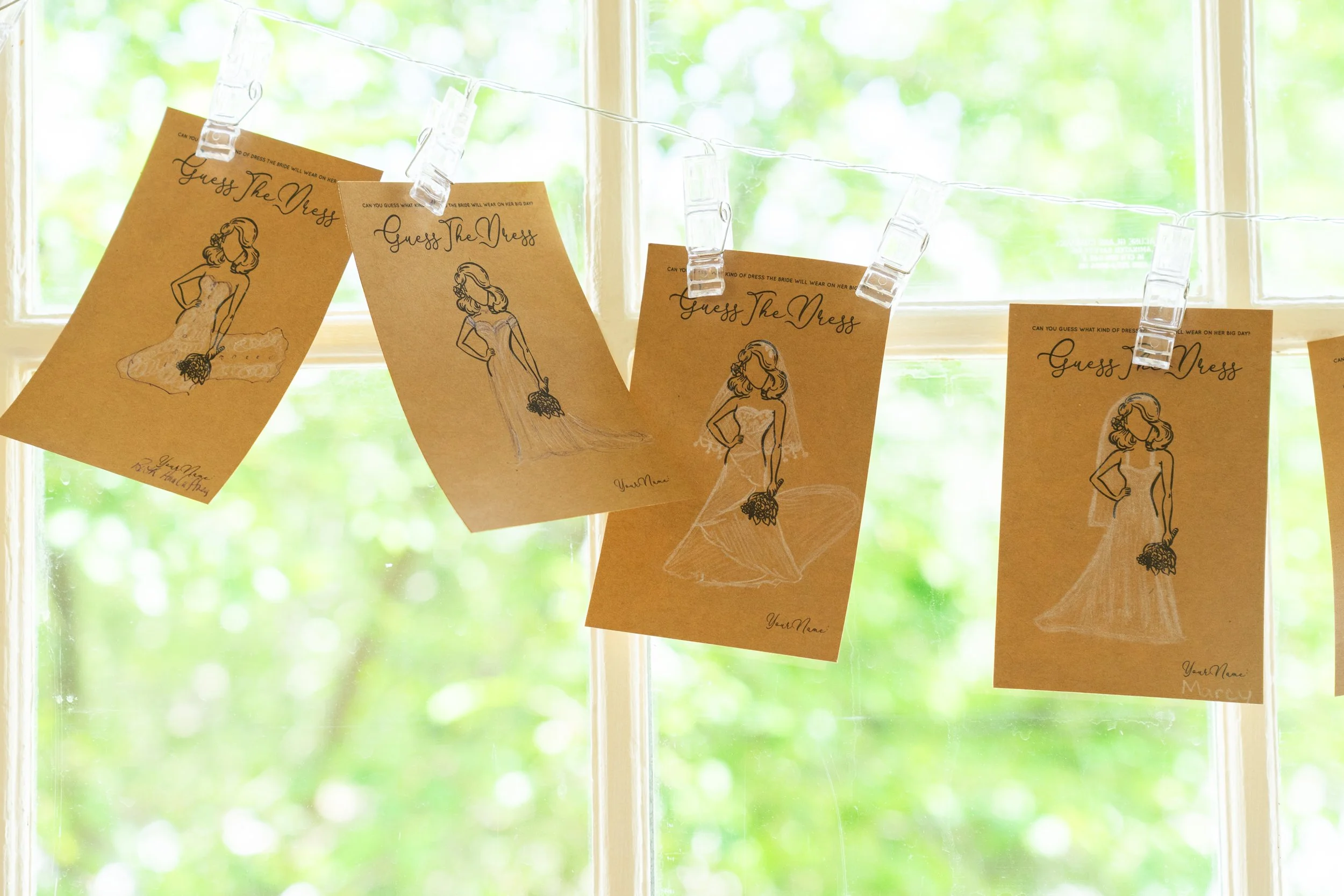 Multiple brown paper cards with a sketch of a woman in a wedding dress, hanging on a string with clothespins against a window with green foliage outside.