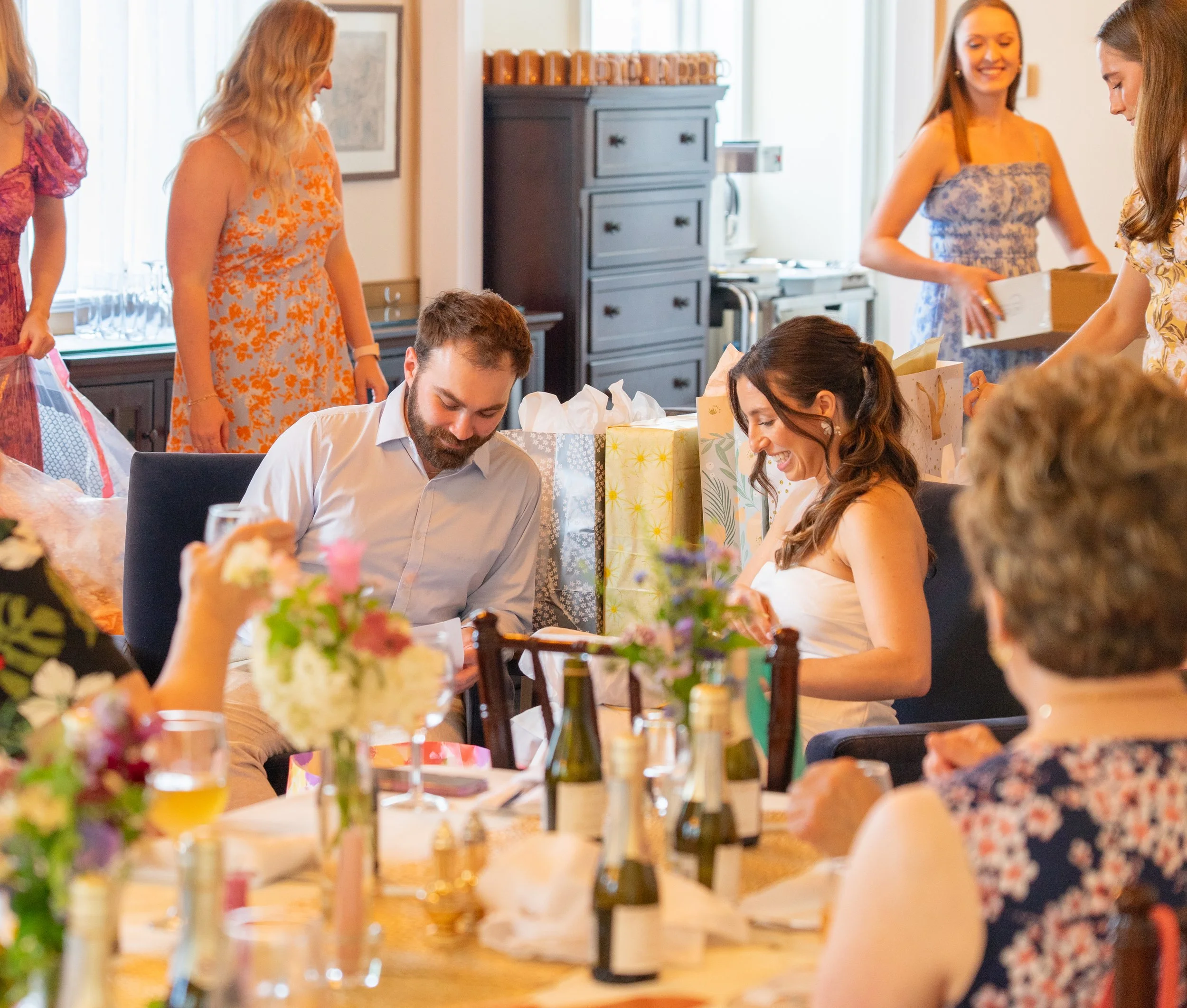 People gathered at a celebration, sitting at a decorated table with flowers and drinks, opening presents, in a bright indoor setting.