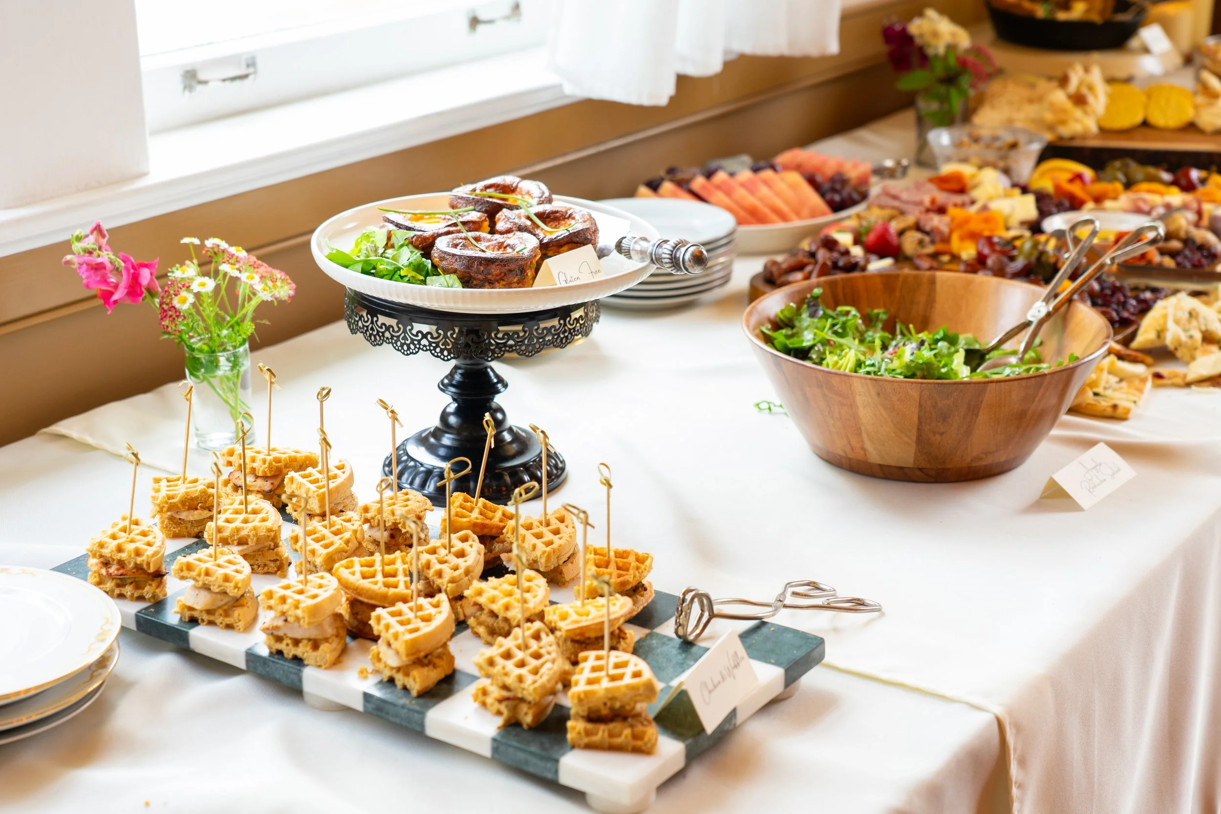Buffet table with waffles, salad, fruit, and various finger foods at a brunch or breakfast gathering.