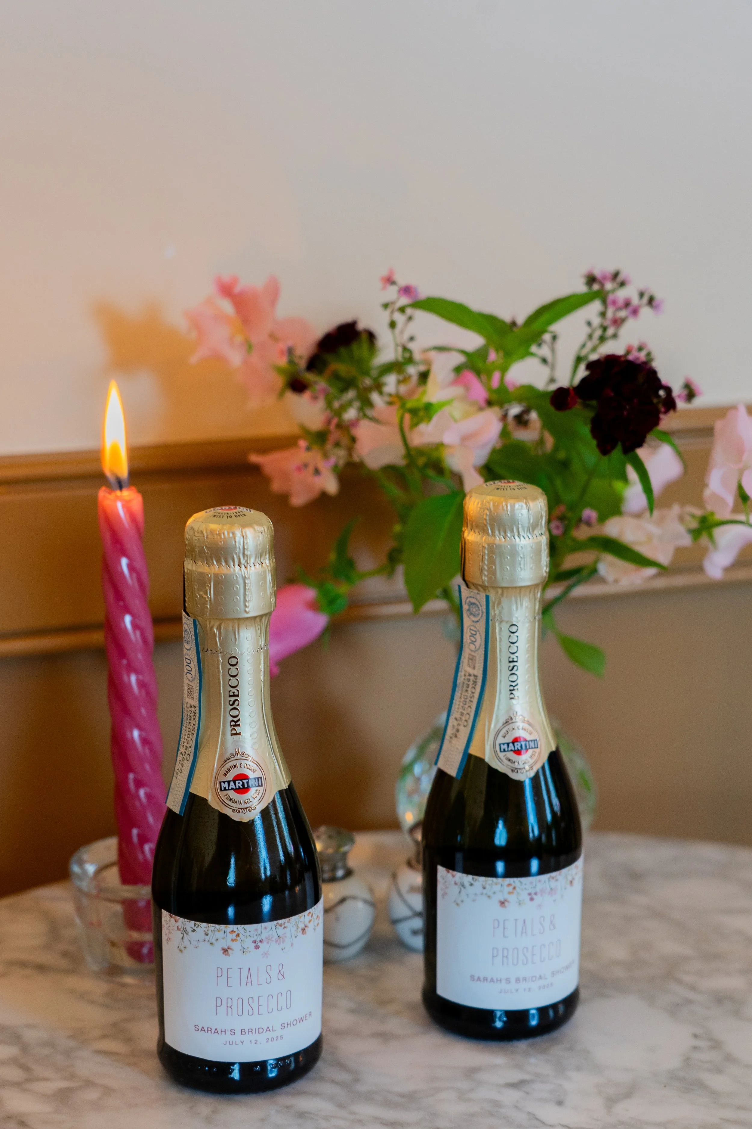Two bottles of petal and prosecco wedding favors with a pink birthday candle and pink flowers in a vase in the background