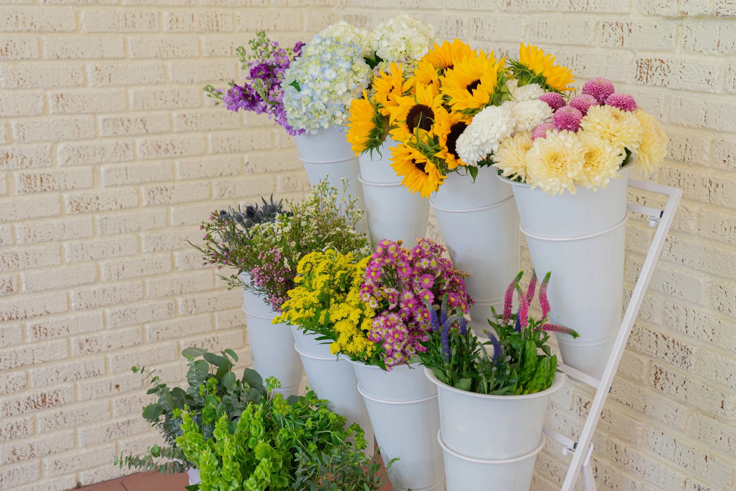 Various colorful flowers in white buckets arranged on a stand against a brick wall.