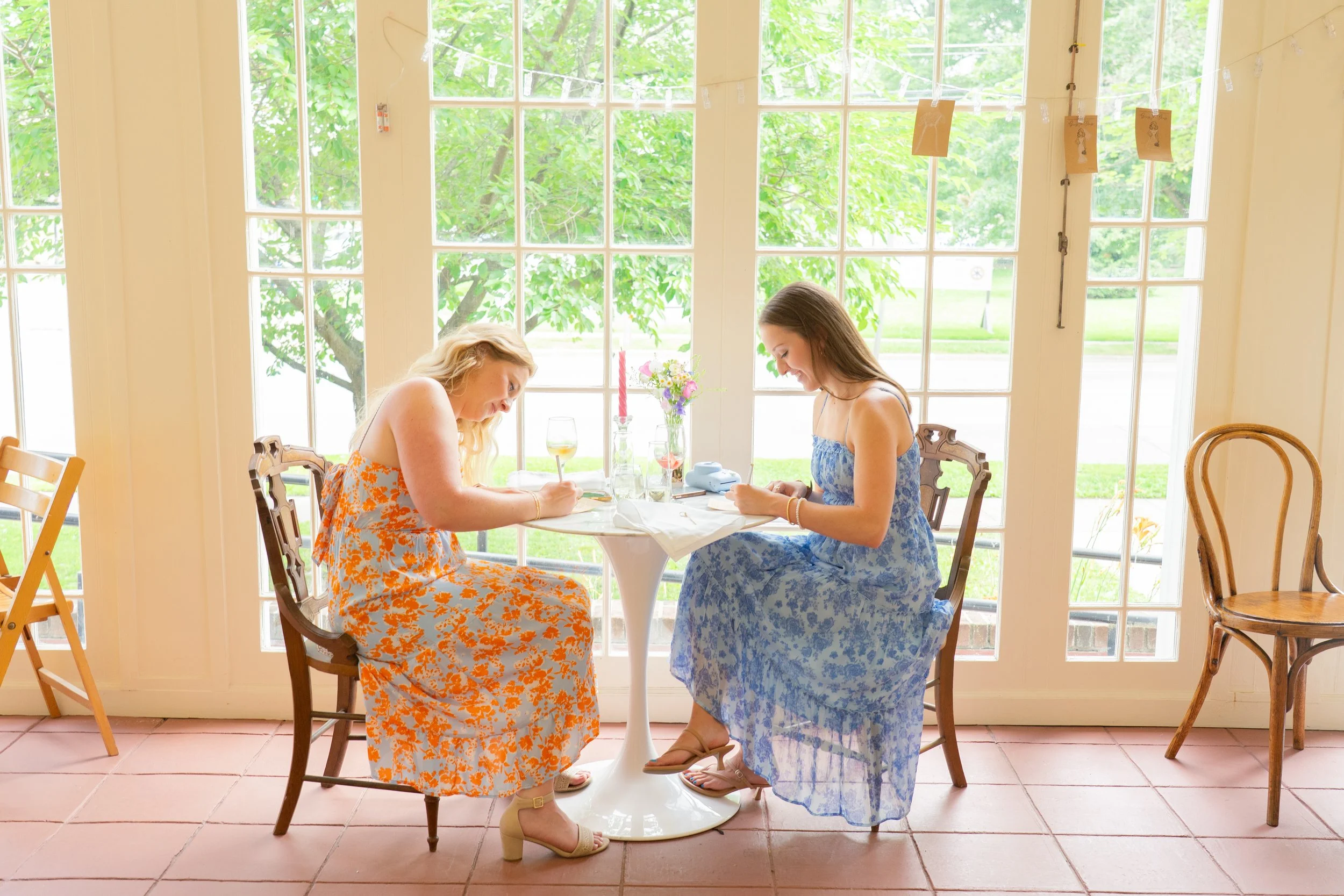 Two women sit at a round table with a white cloth, writing on cards in a bright room with large glass doors. One woman has blonde hair and wears an orange floral dress, the other has brown hair and wears a blue floral dress. There are wine glasses, a