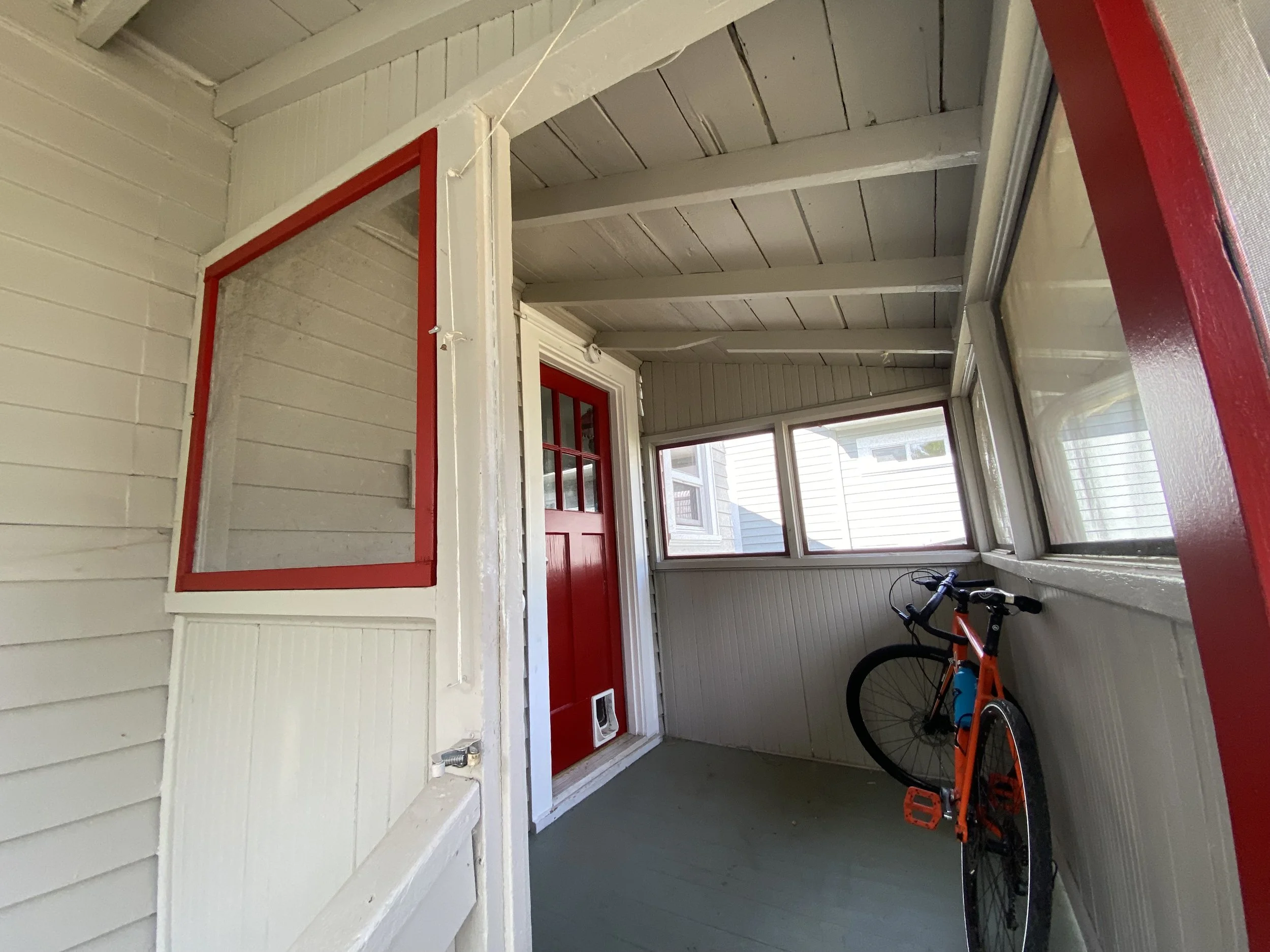 A small enclosed porch with white painted wood walls and ceiling, red trim around windows and door, a bicycle with orange and black frame parked inside, and multiple windows letting in natural light.