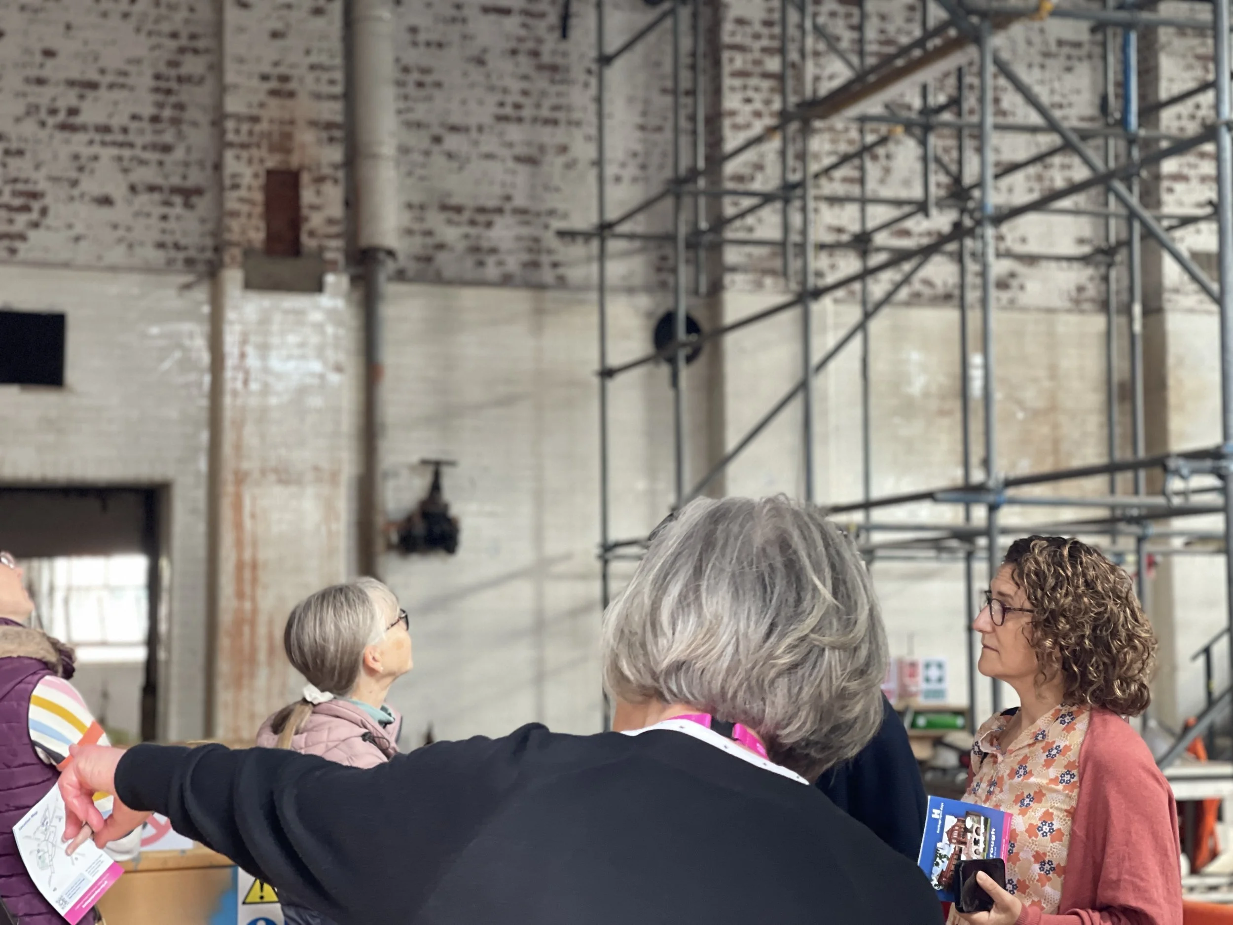 A group of women engaged in a conversation inside an industrial-style building with exposed brick walls and metal scaffolding.