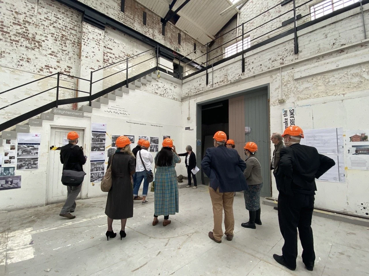 Group of people wearing orange safety helmets inside an industrial-style building with exposed brick and white walls, viewing displayed photos and information.