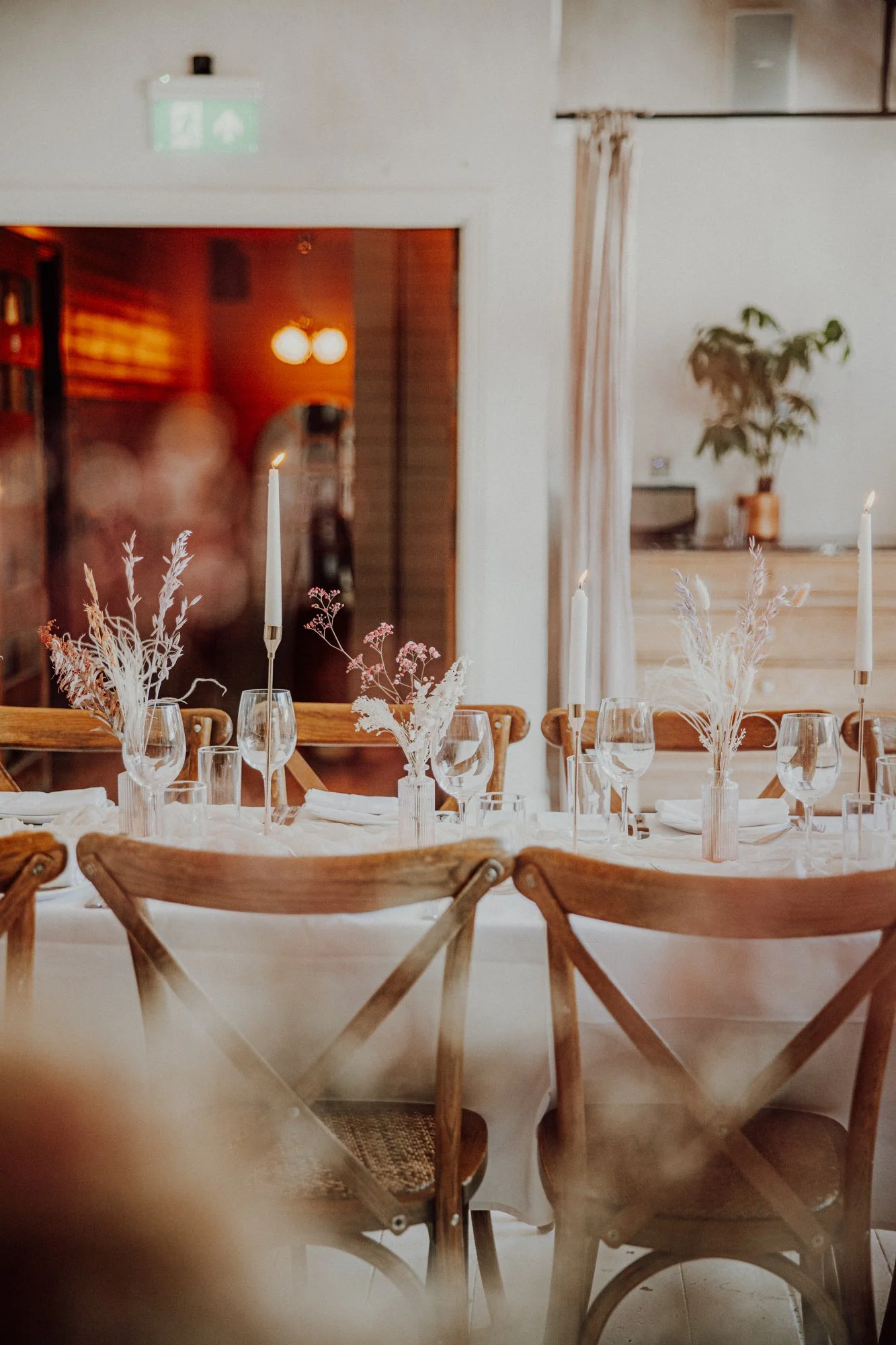 A decorated dining table with white tablecloth, wooden chairs, tall white candles, glassware, and dried floral arrangements, in a cozy indoor setting with warm lighting.