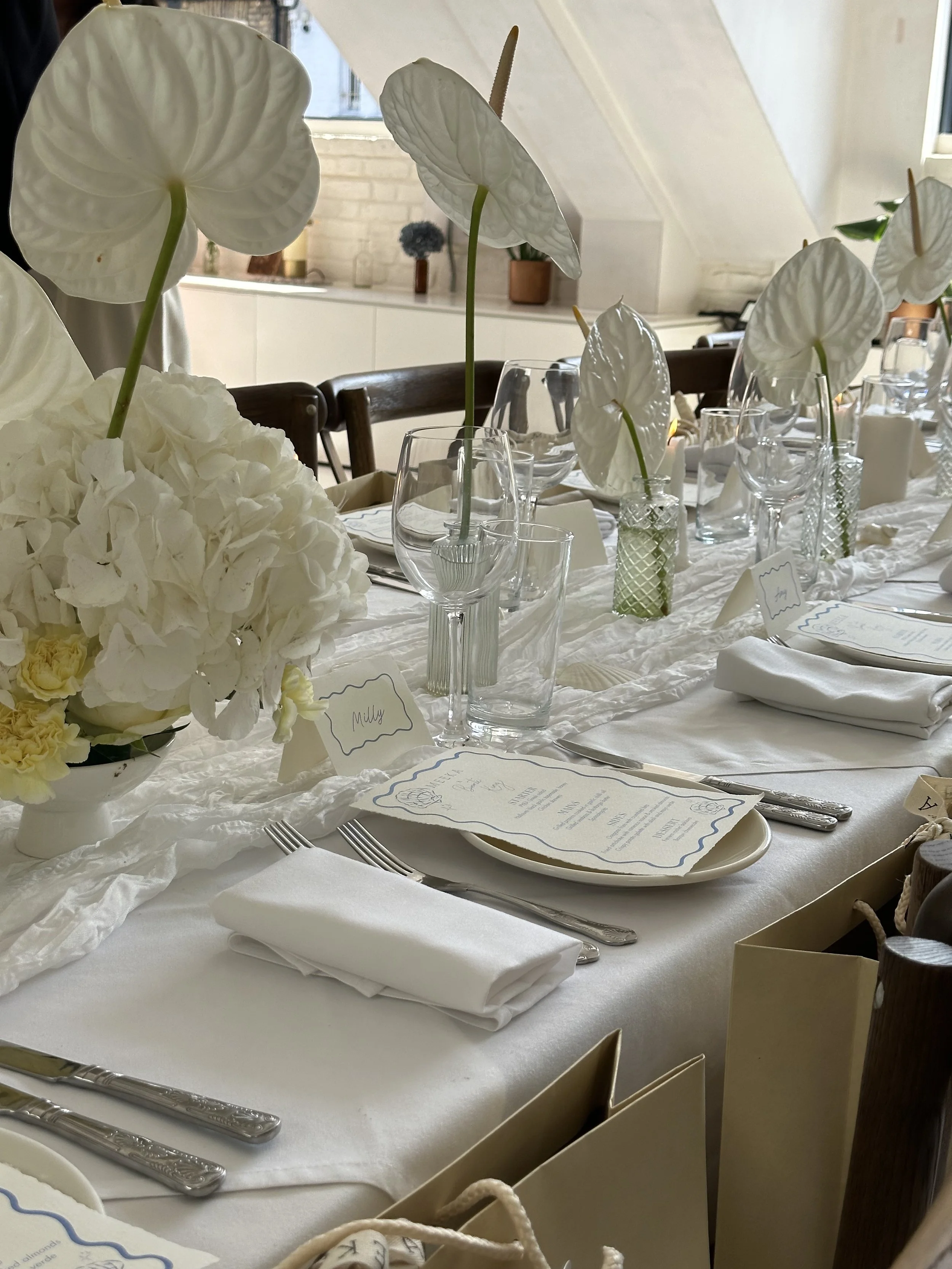 A dining table set with white flowers, calligraphy menus, white napkins, glassware, and cutlery, in a bright room with windows and white walls.