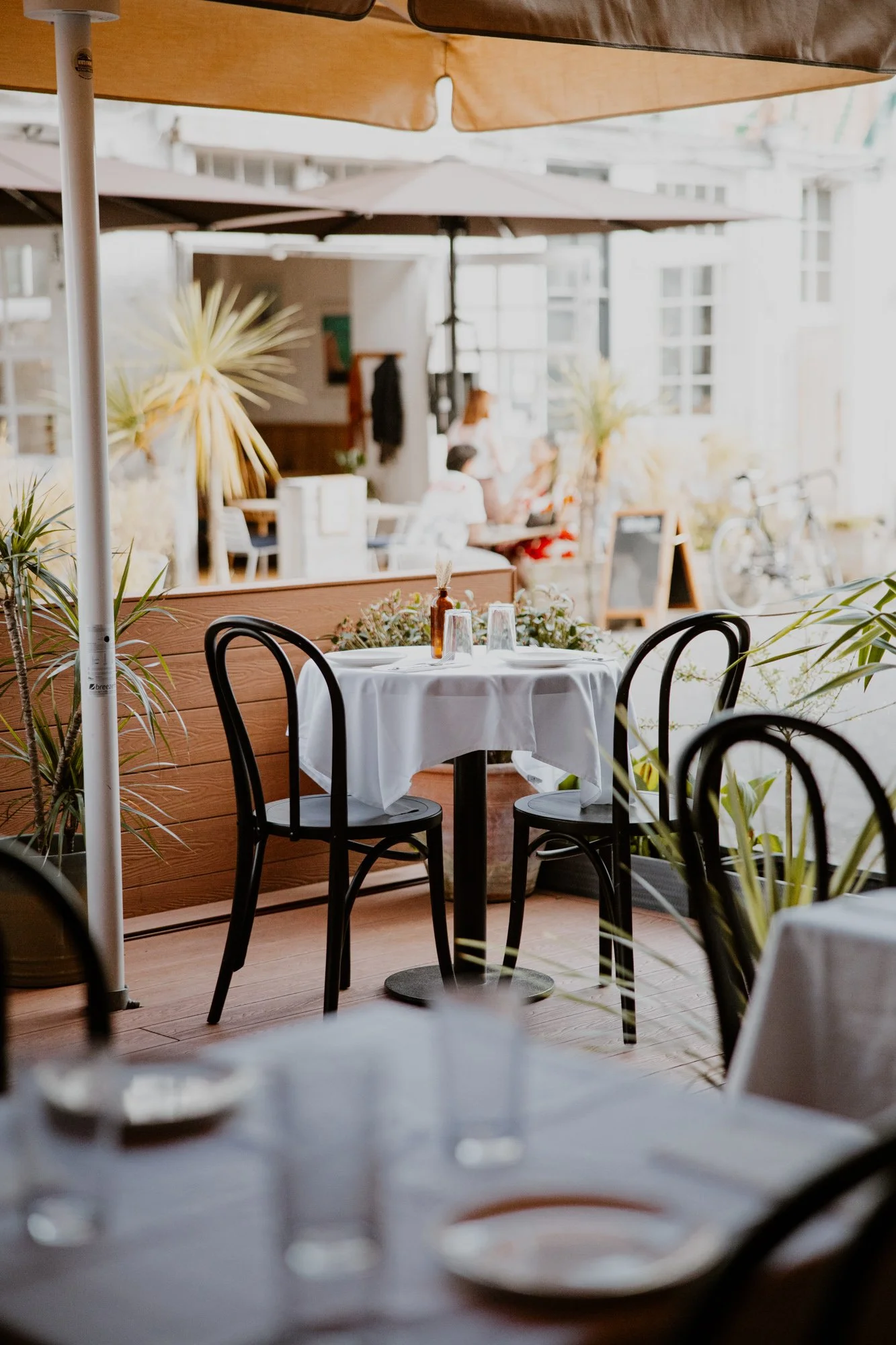 Empty outdoor cafe table with white tablecloth, glasses, and a small vase, surrounded by black chairs, with plants and other tables in the background.