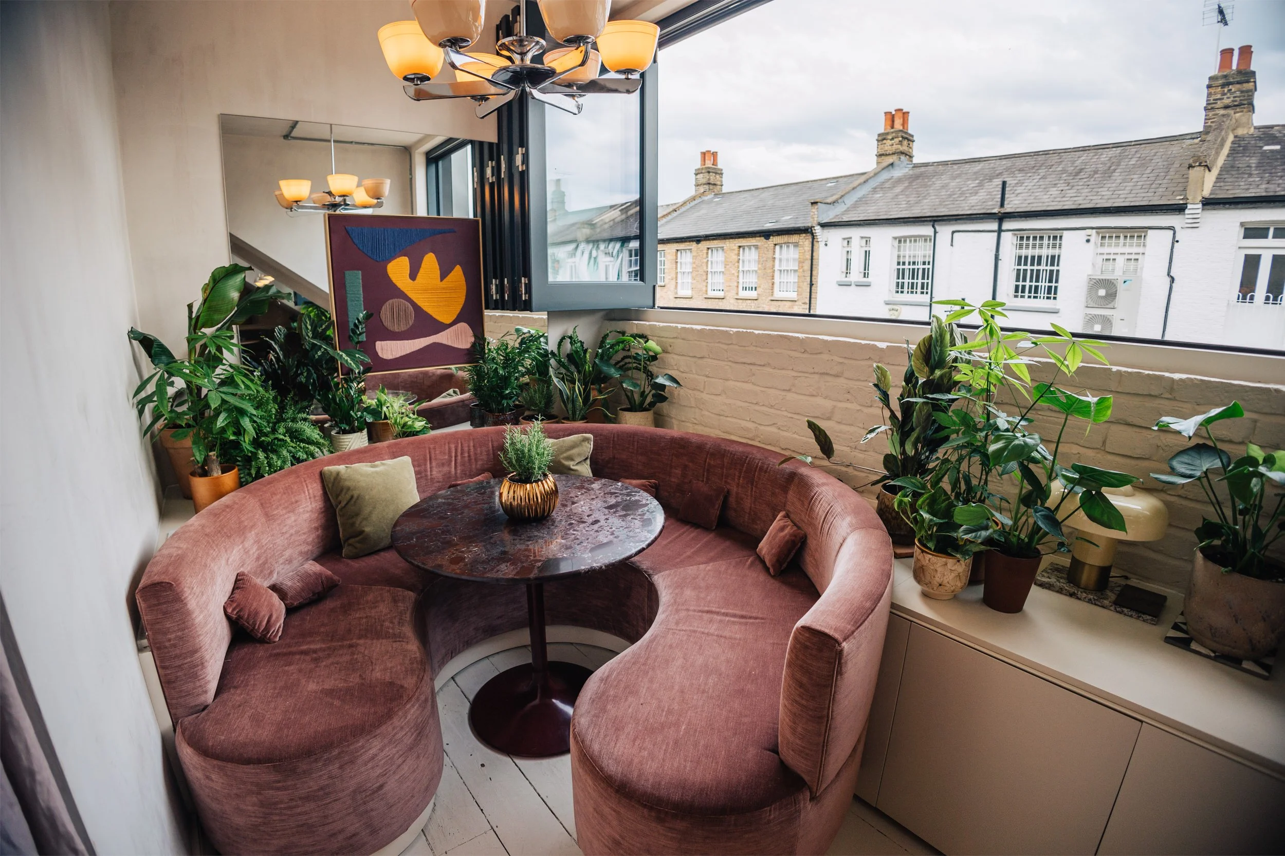 A cozy corner with a curved, pink velvet banquette, a round marble table with a small potted plant, and several potted plants along a white brick wall next to a large window showing neighboring rooftops.