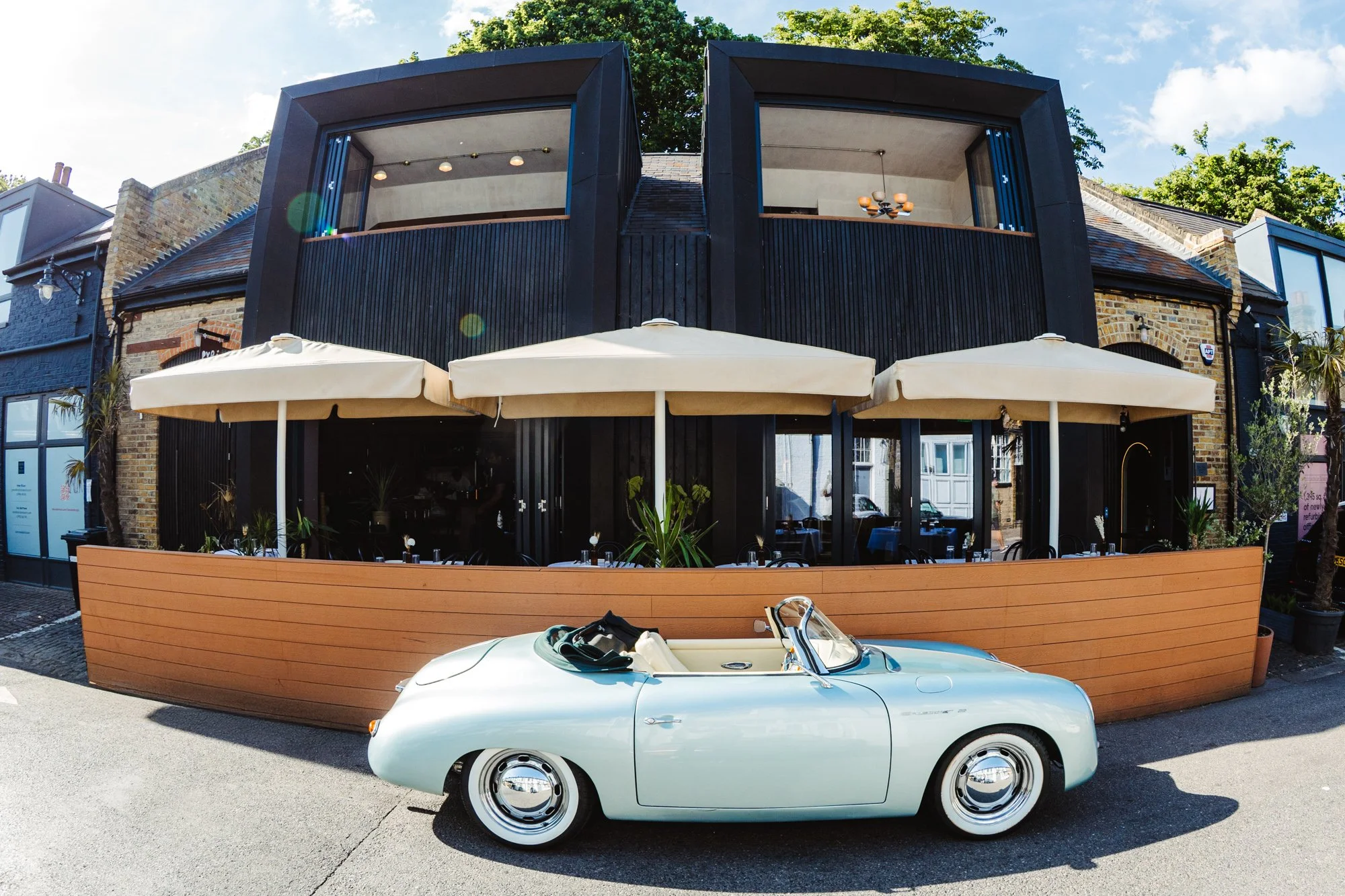 A vintage white convertible car parked in front of a modern restaurant with outdoor seating and large umbrellas.