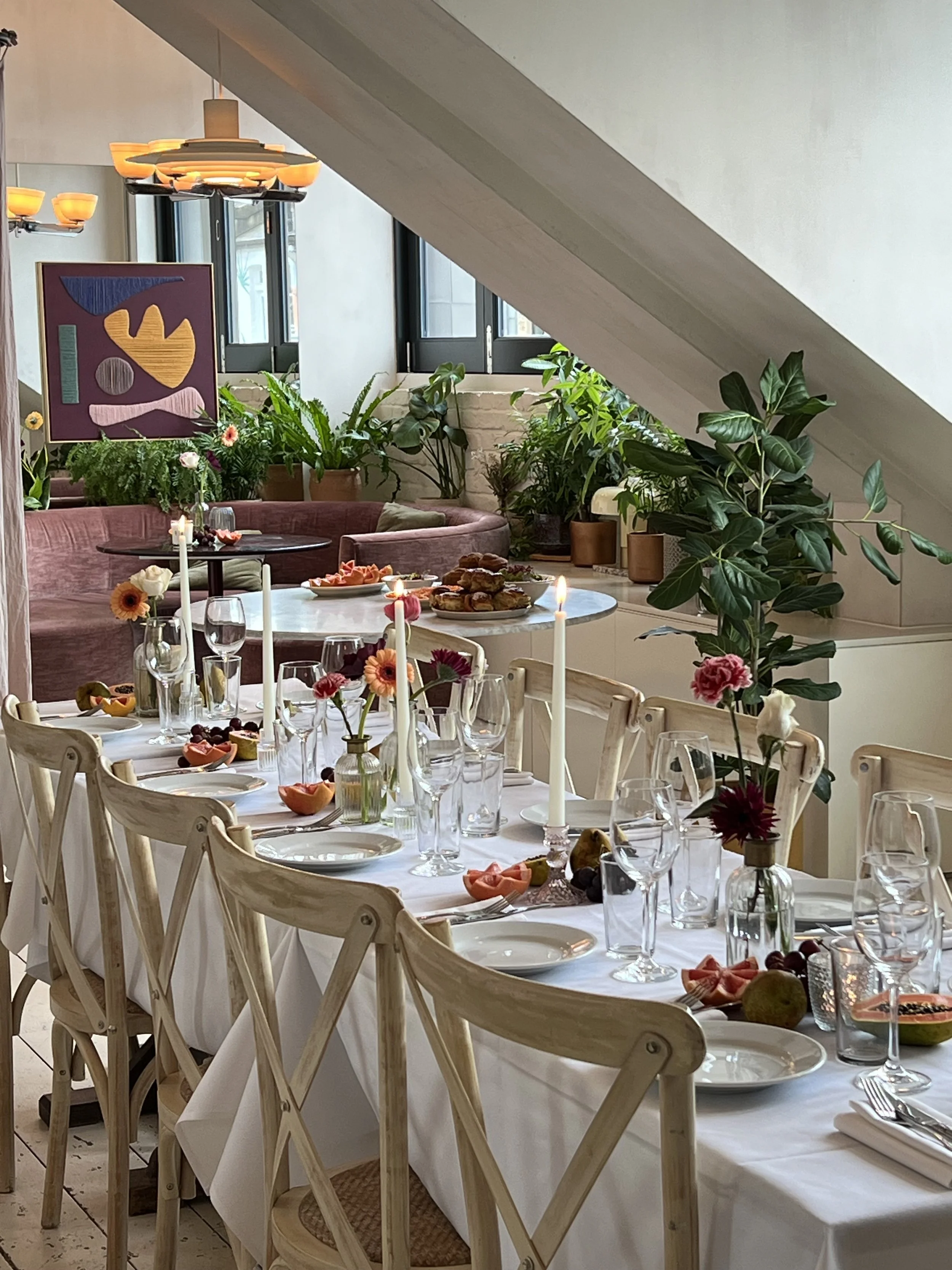 Elegant dining table set for a meal with candles, flowers, and various foods in a bright, modern room with plants and abstract art.