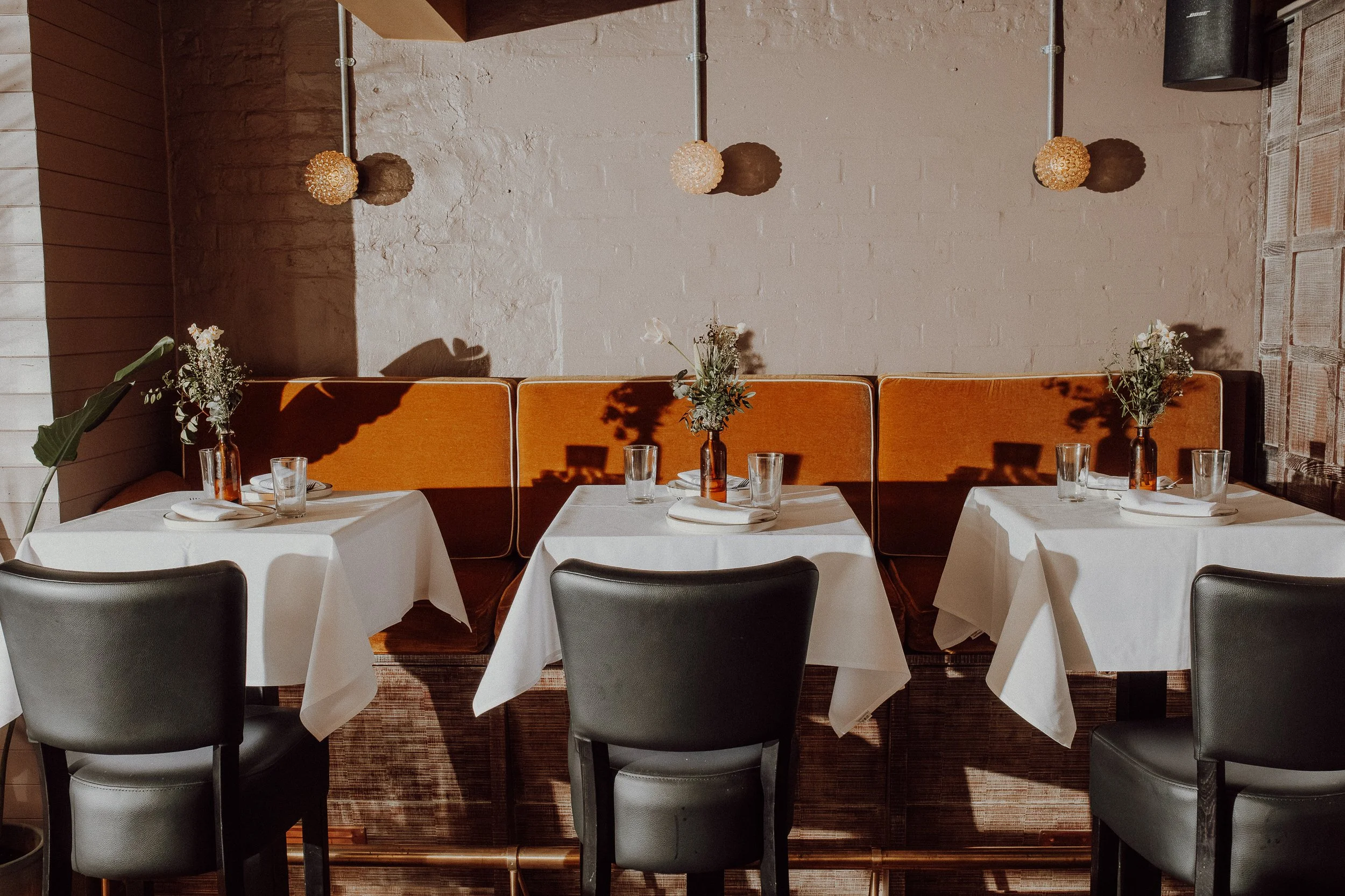 Three tables with white tablecloths set with glasses, plates, and small flower arrangements in amber vases in a restaurant with dim lighting and a cream brick wall.