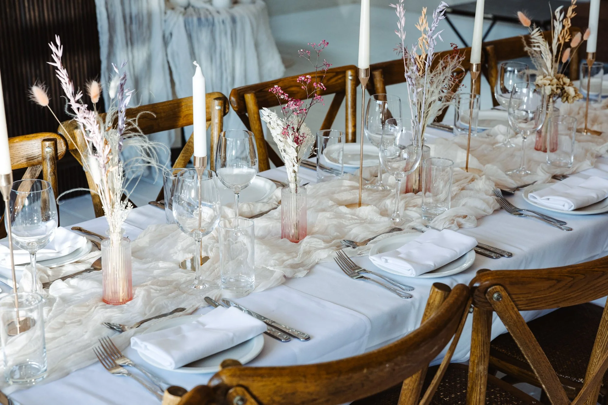 Elegant dining table set with white tablecloth, white napkins, clear glassware, silverware, and decorative pink and purple vases with dried flowers, lit by tall white candles in candleholders.