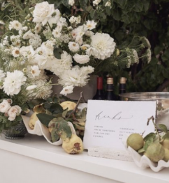 Elegant display with white flowers, wine bottles, green pears, and a menu card on a white surface.