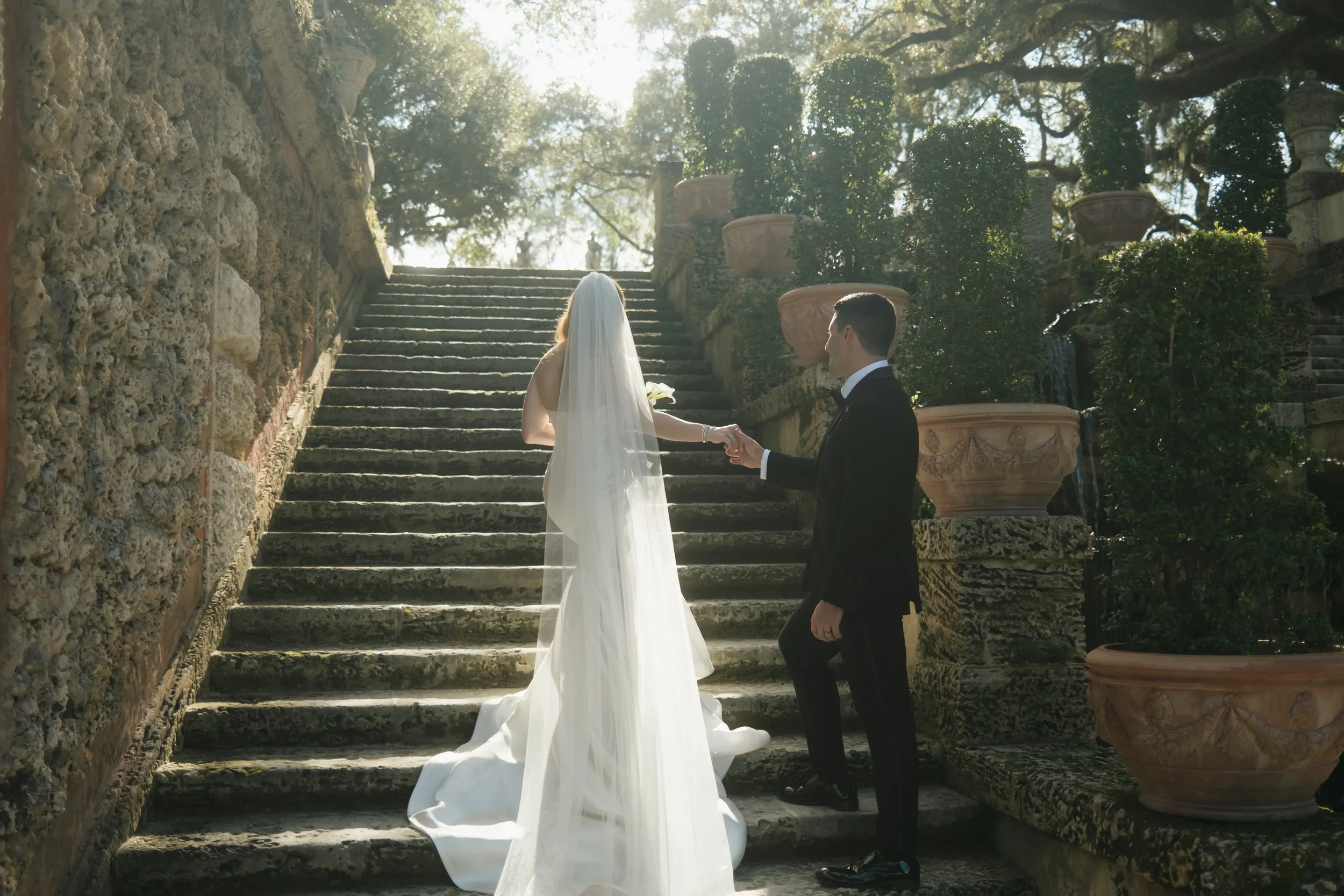 A bride and groom holding hands in an outdoor garden setting, standing on stone steps with trees and large plant pots in the background, sunlight filtering through the branches.