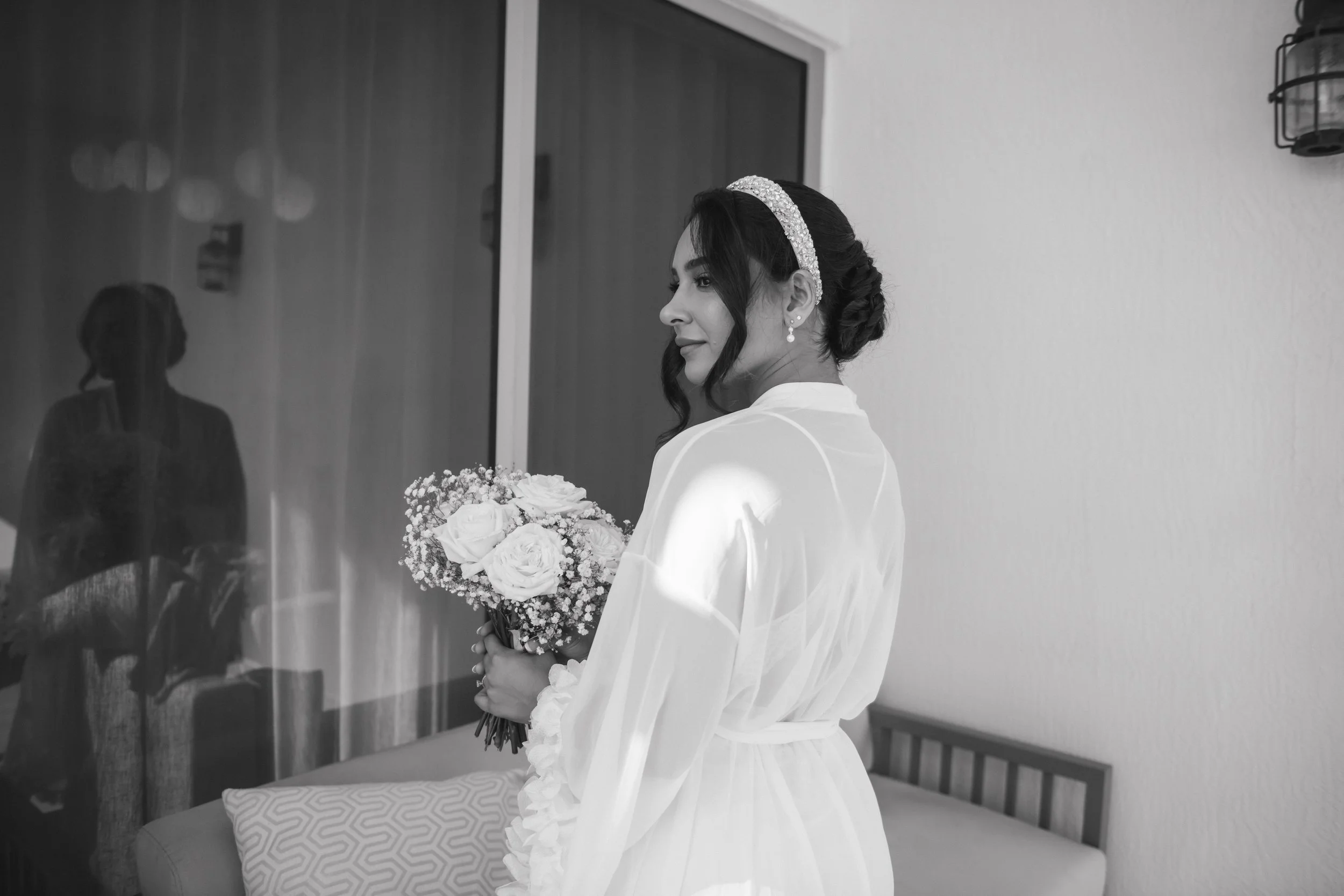 Bridal woman in white robe holding bouquet of flowers inside a room with sofa and wall-mounted lamp, reflected in mirror.